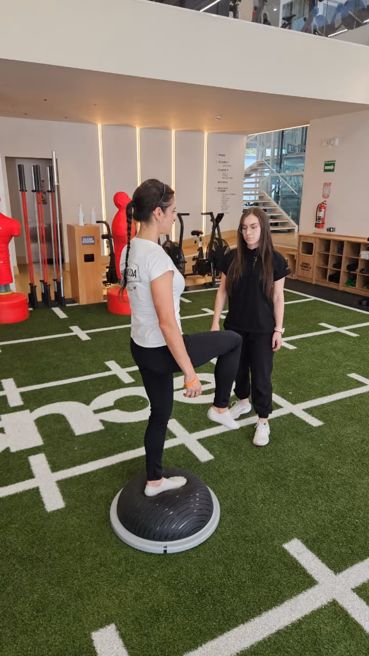 Una mujer de pie en un dispositivo de equilibrio mientras otra la observa en un gimnasio.
