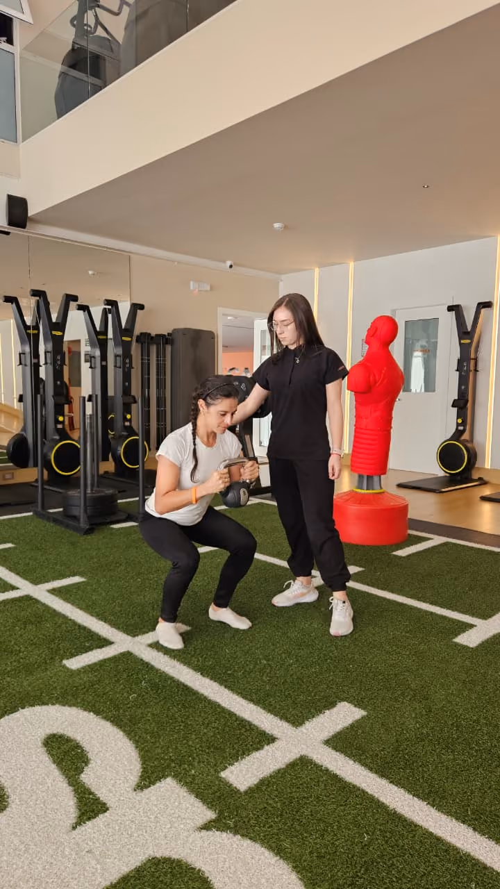 Mujer con pesa haciendo sentadillas asistida por entrenadora en gimnasio con suelo de césped artificial.