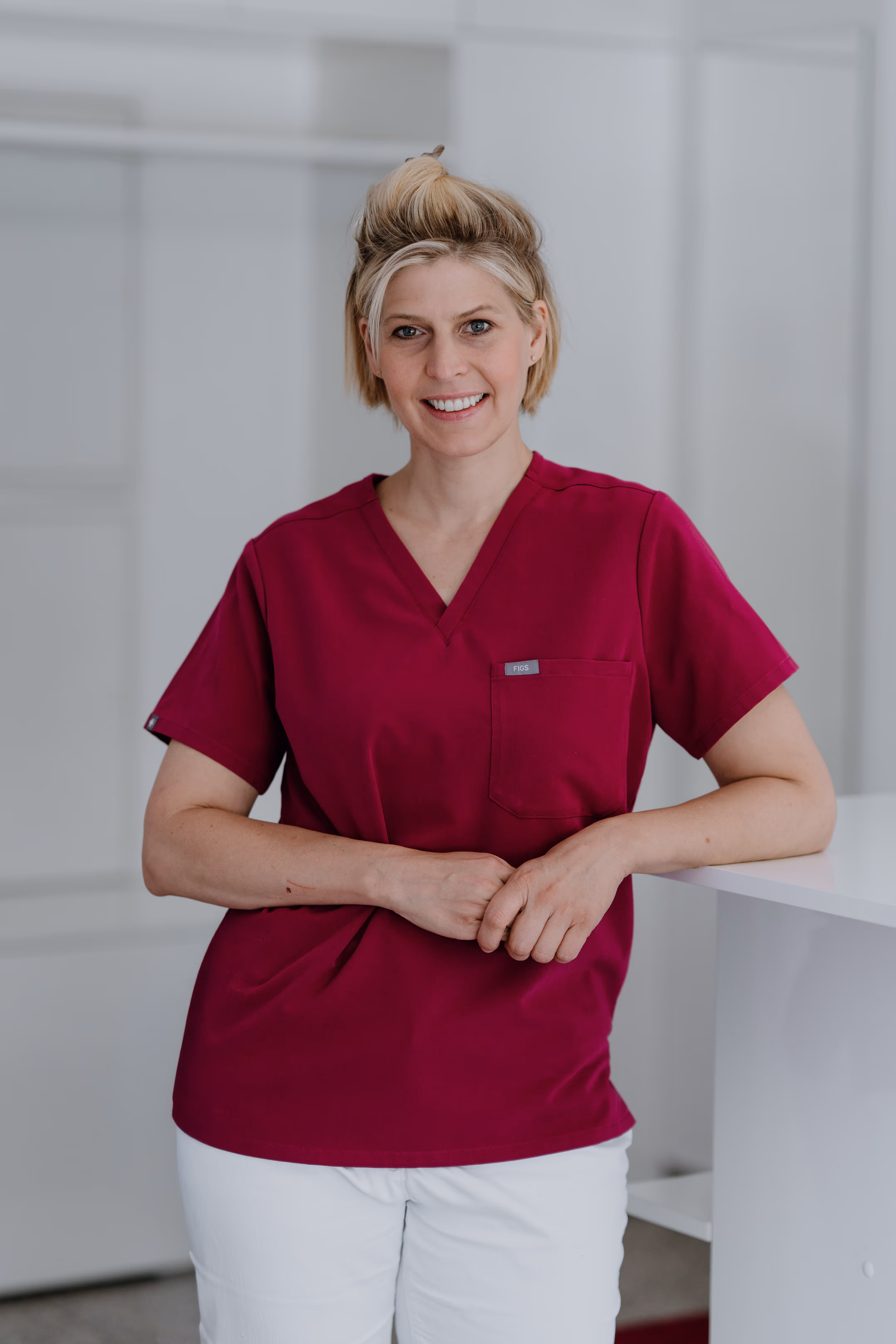 Smiling woman in a red medical scrub leans against a white counter in a bright setting.