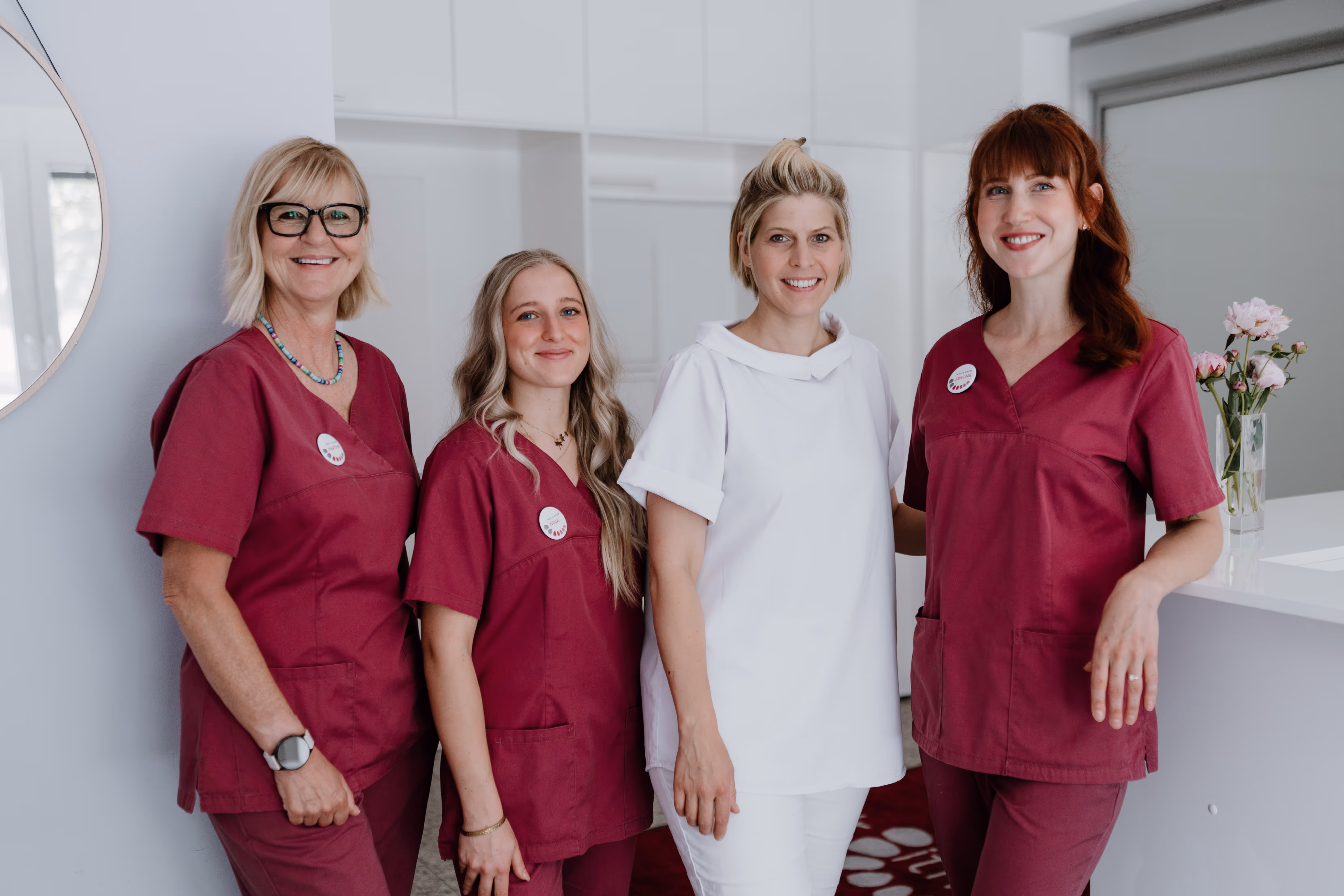Zwei lächelnde Frauen in weißen Kitteln stehen vor einer weißen Wand mit Bildern rechts.