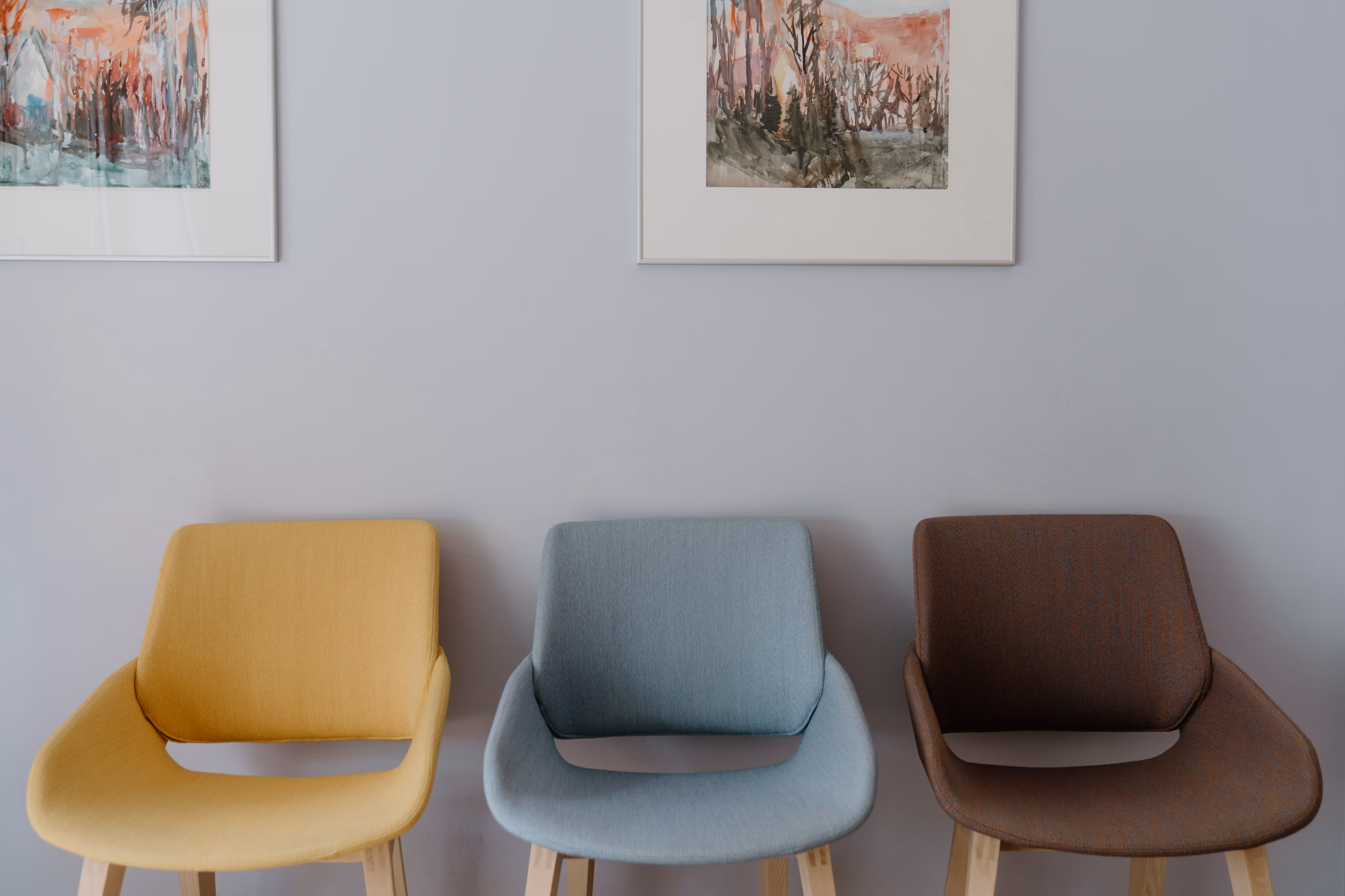 Drei moderne Stühle in Gelb, Blau und Braun vor einer hellgrauen Wand mit zwei abstrakten Gemälden.
