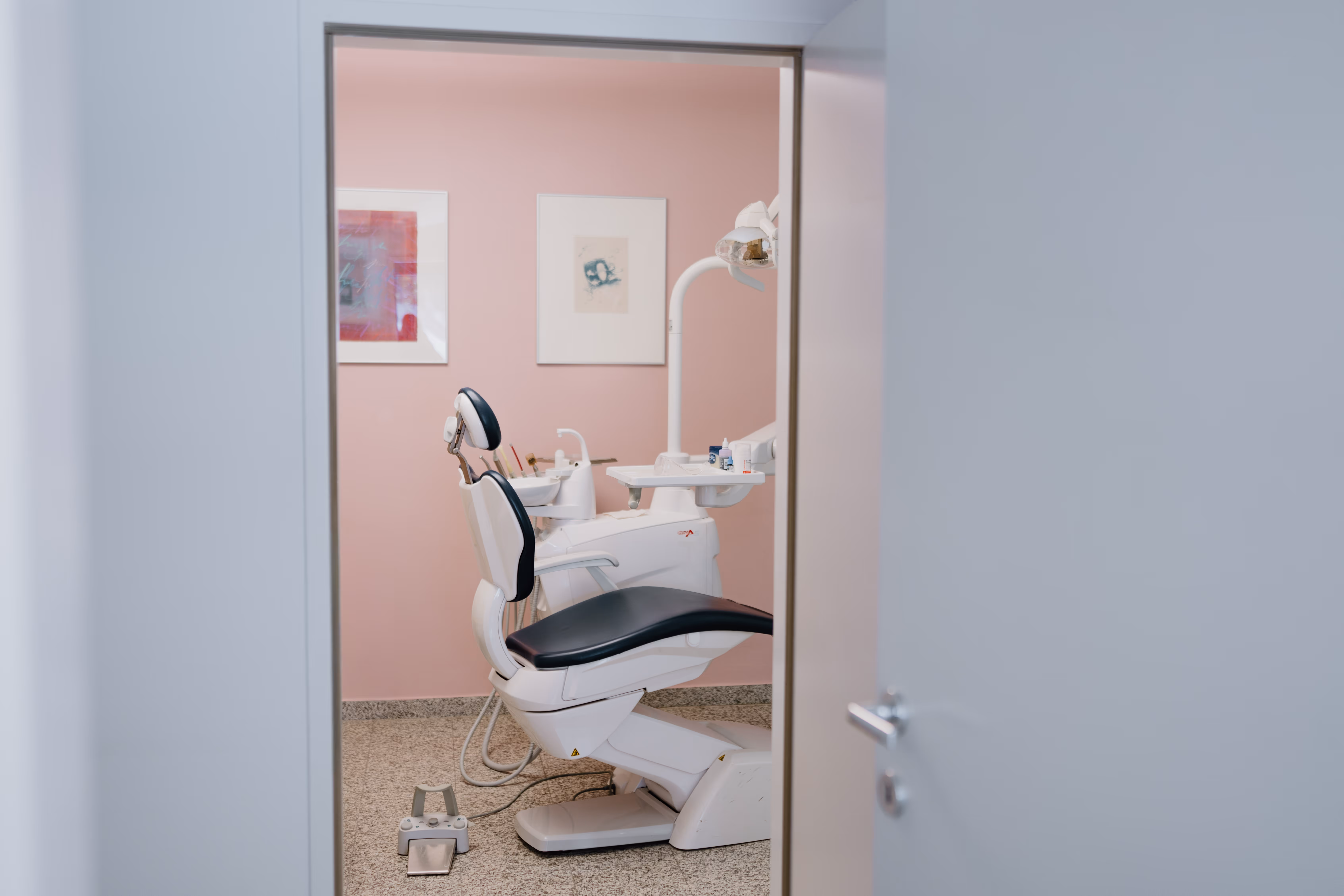 Blick durch offene Tür auf einen zahnärztlichen Behandlungsstuhl in einem Raum mit rosa Wänden.