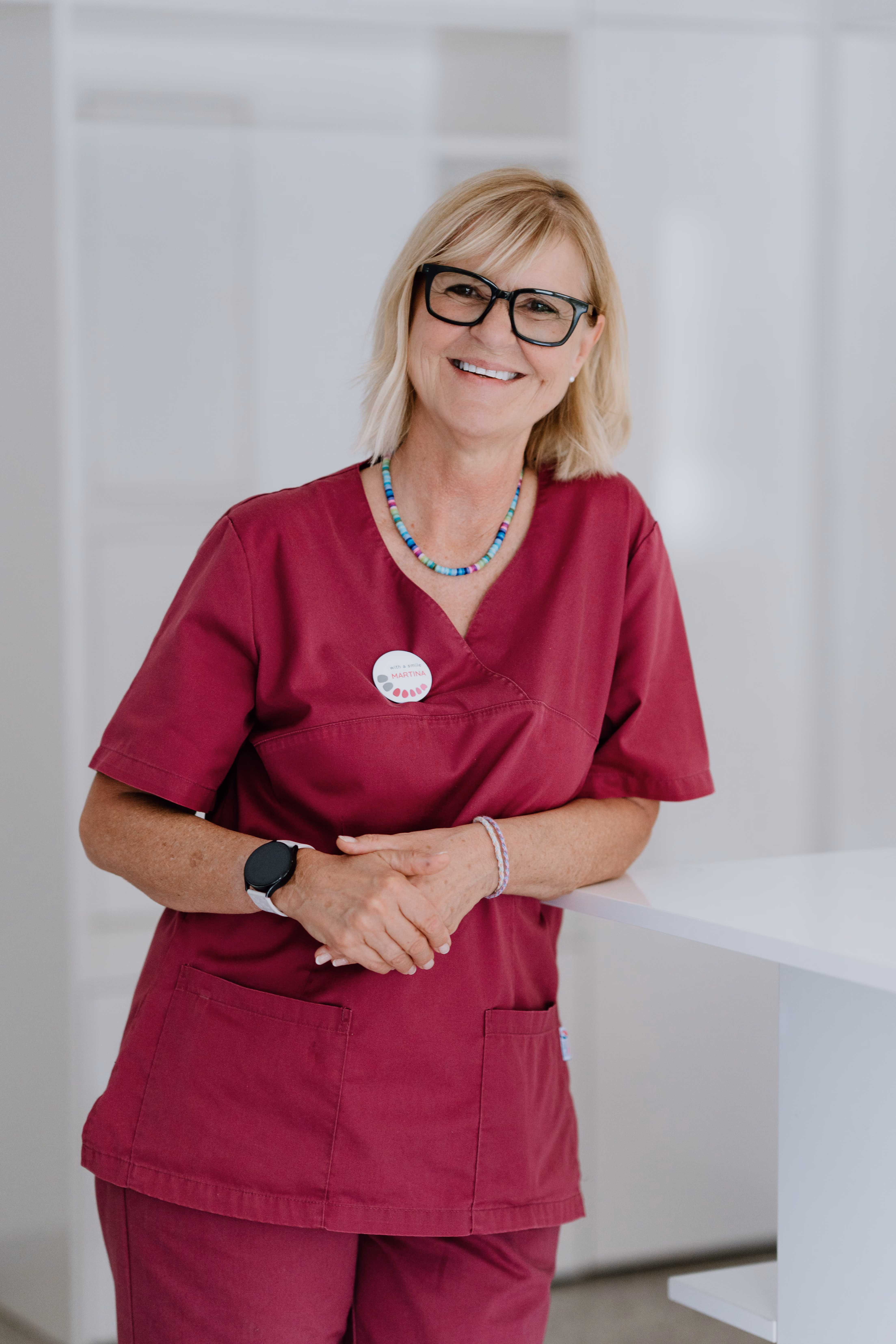 Lächelnde Frau mit Brille und bordeauxroter Arbeitskleidung steht in hellem Raum.