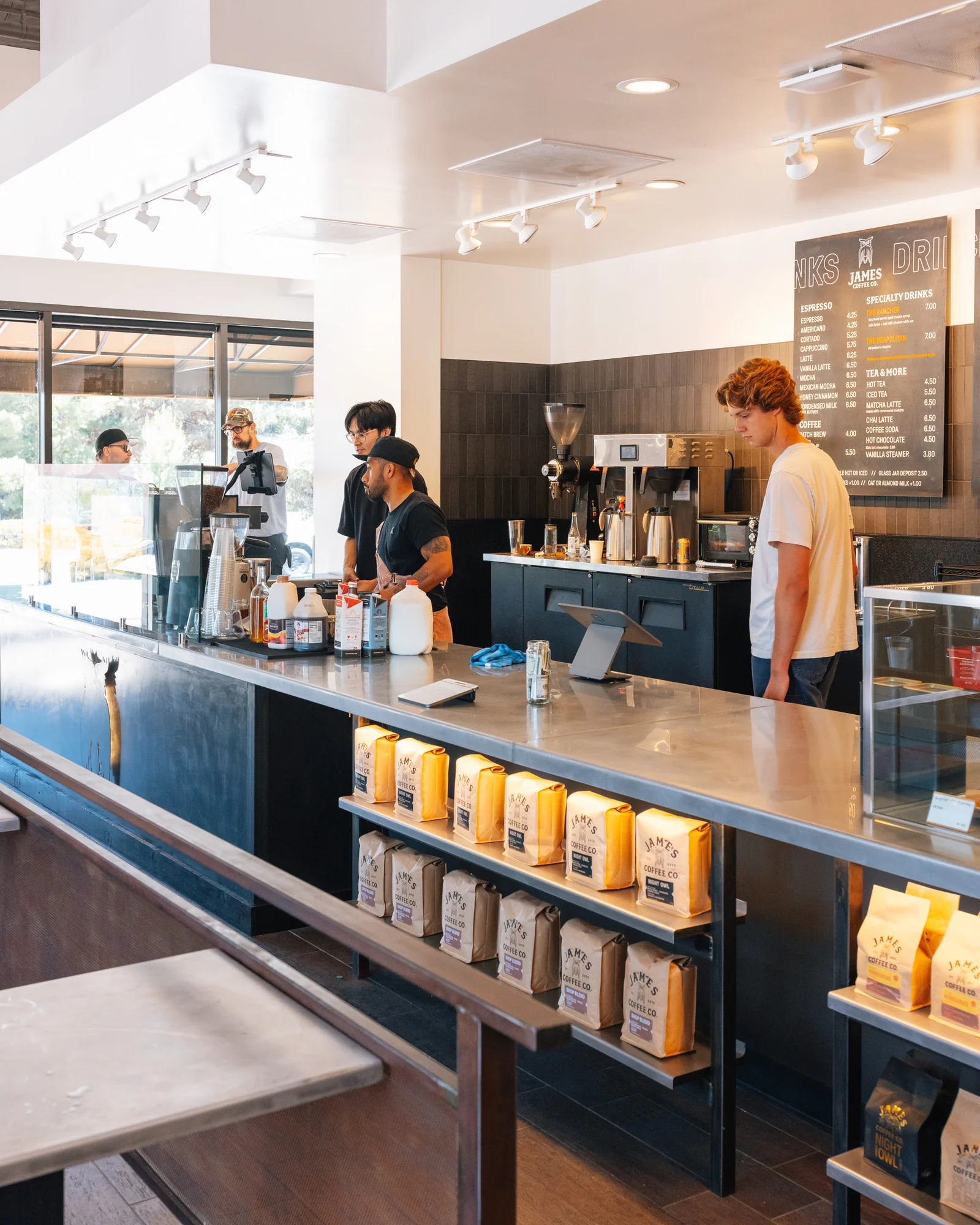 Three baristas behind counter in coffee shop with coffee bags displayed under counter and menu on wall.