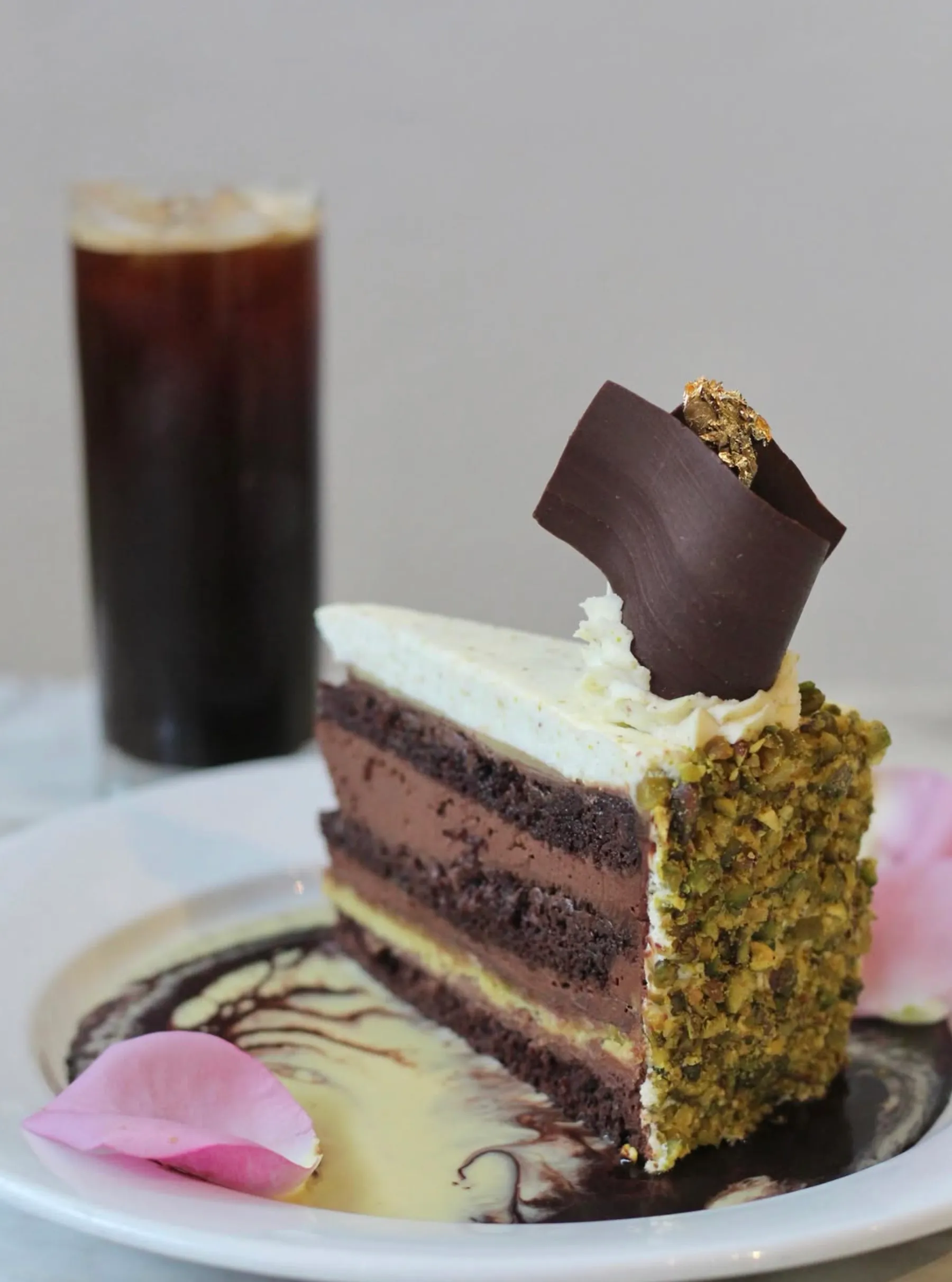 Slice of layered chocolate and cream cake with pistachio crust and chocolate curl, served with sauce and rose petals.