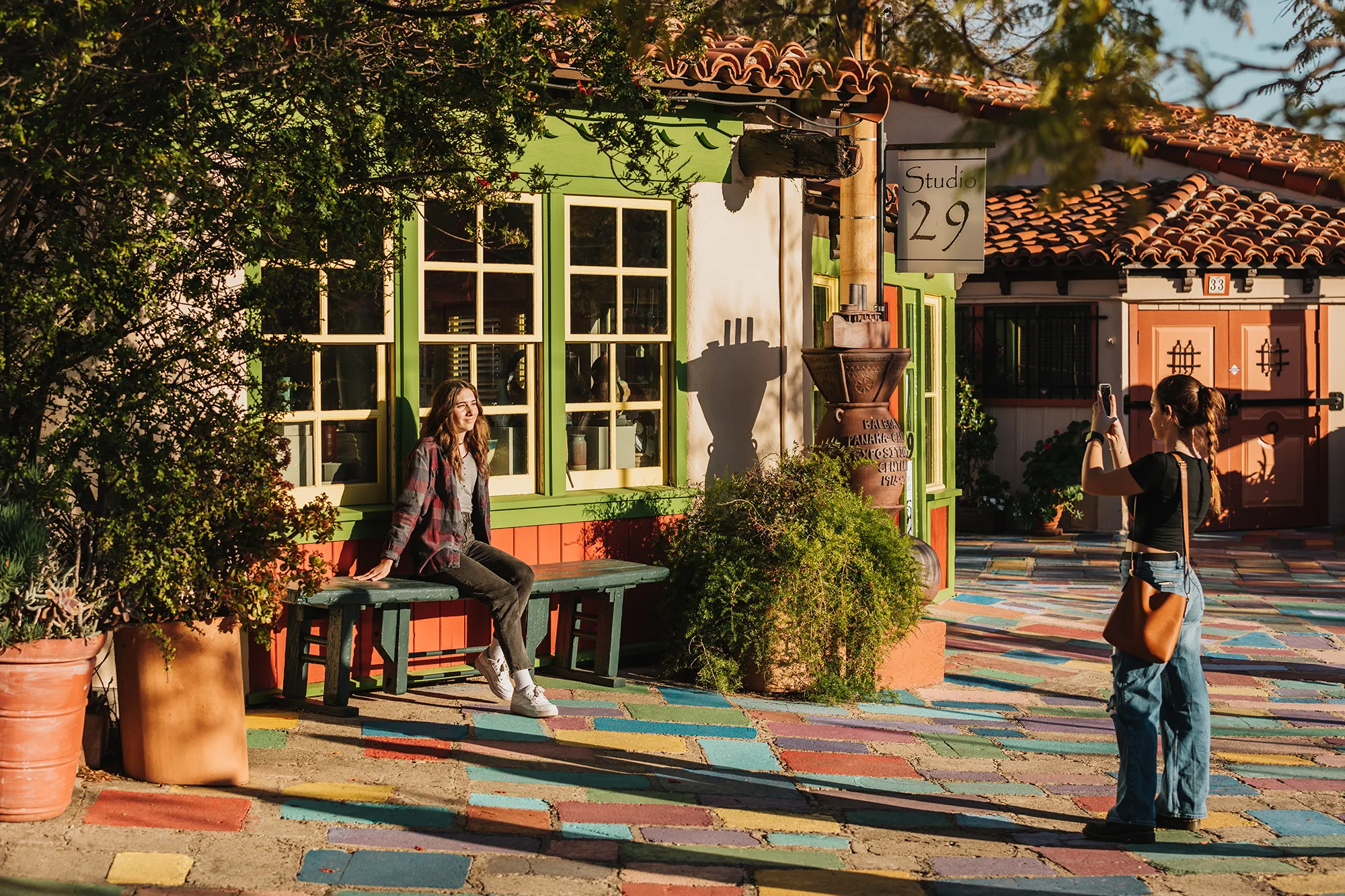 Woman sitting on a bench in front of a colorful building while another woman takes her photo outdoors.