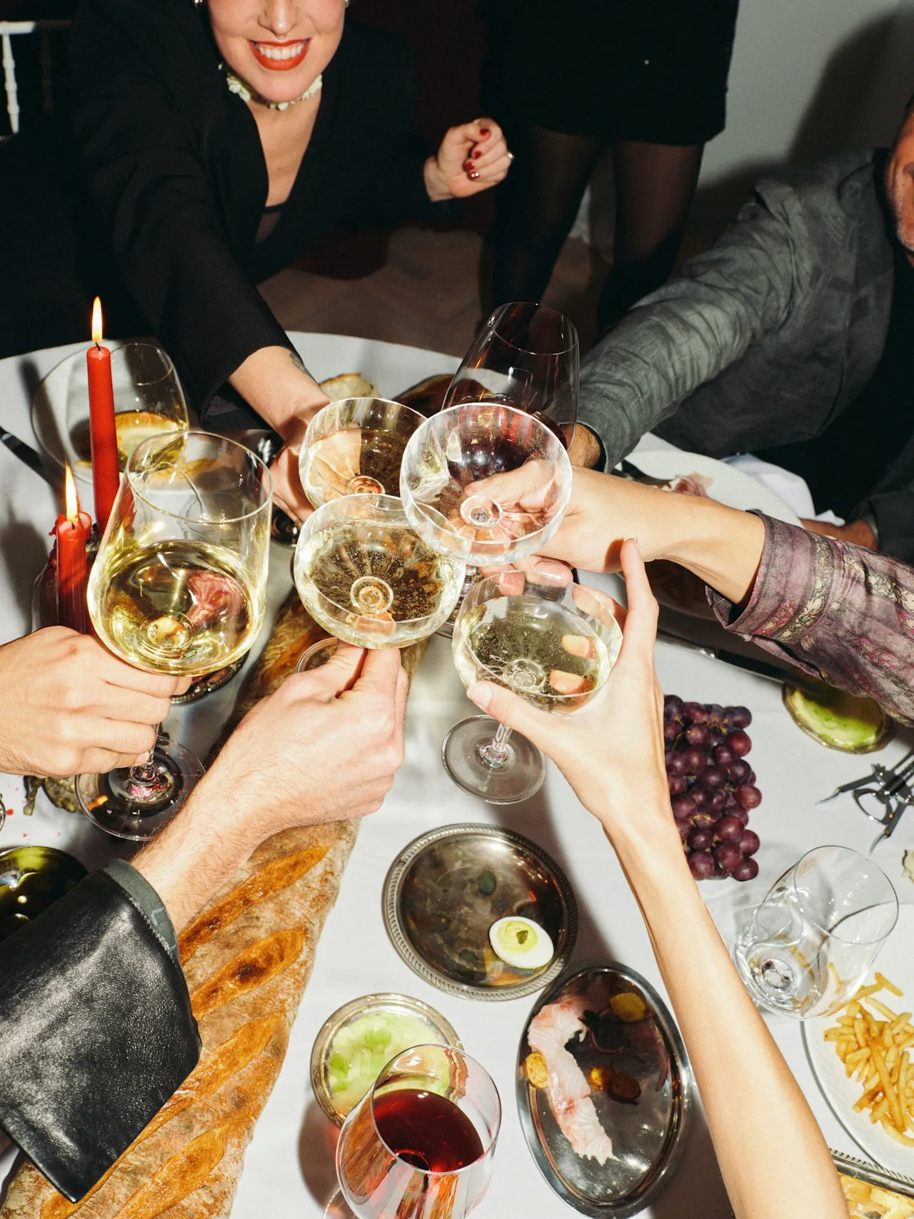 People raising glasses of white and red wine for a toast over a table with food and candles.