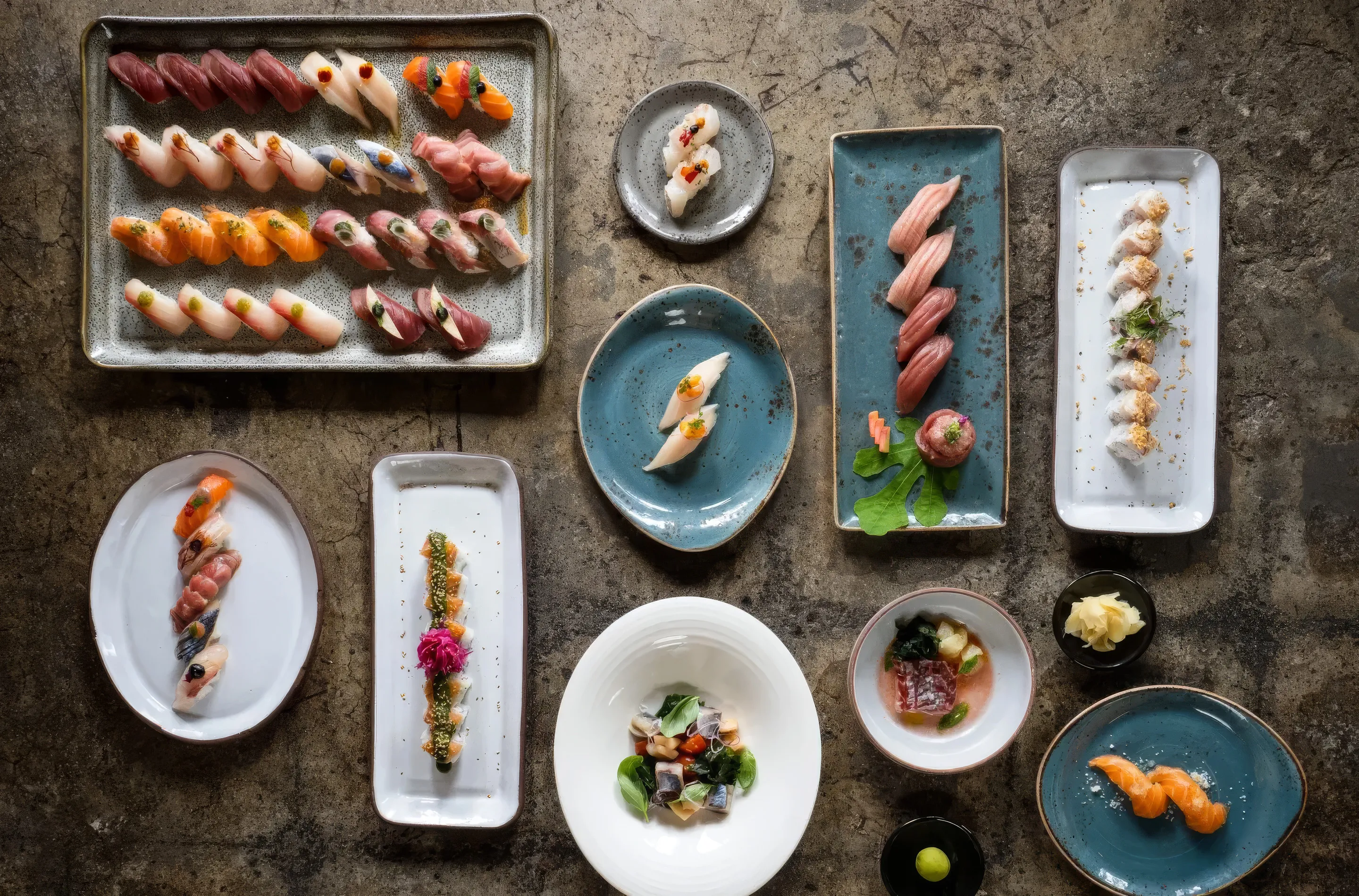 Variety of sushi and sashimi arranged on multiple ceramic plates on a rustic stone surface.