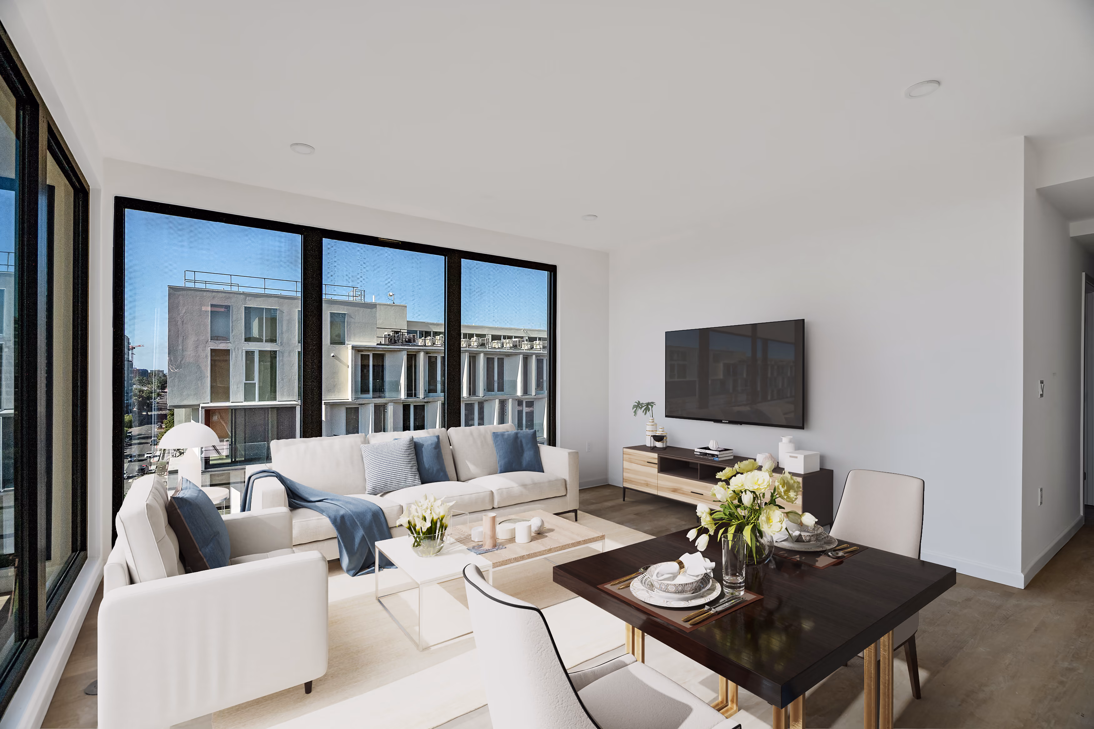 Modern living room with white sofa, armchair, large windows, TV, and a dining table with flowers.