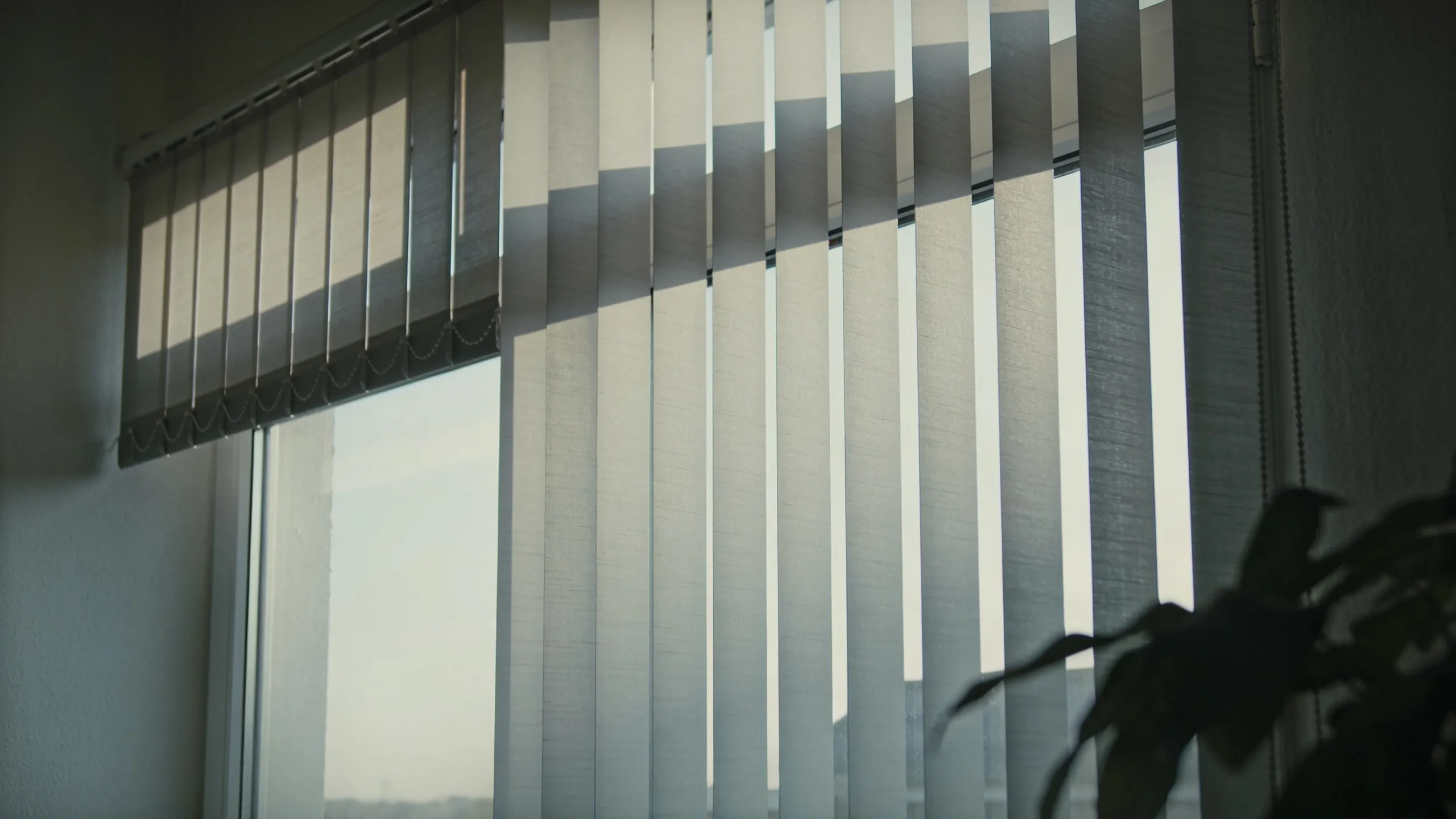 Beige vertikale Lamellenvorhänge an einem Fenster mit Licht und Schatten, Zimmerpflanze im Vordergrund.