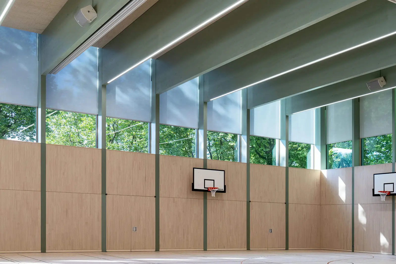 Sporthalle mit hohem Fensterband, an dem eine Reihe heller Rollos auf gleicher Höhe halb herabgelassen ist.
