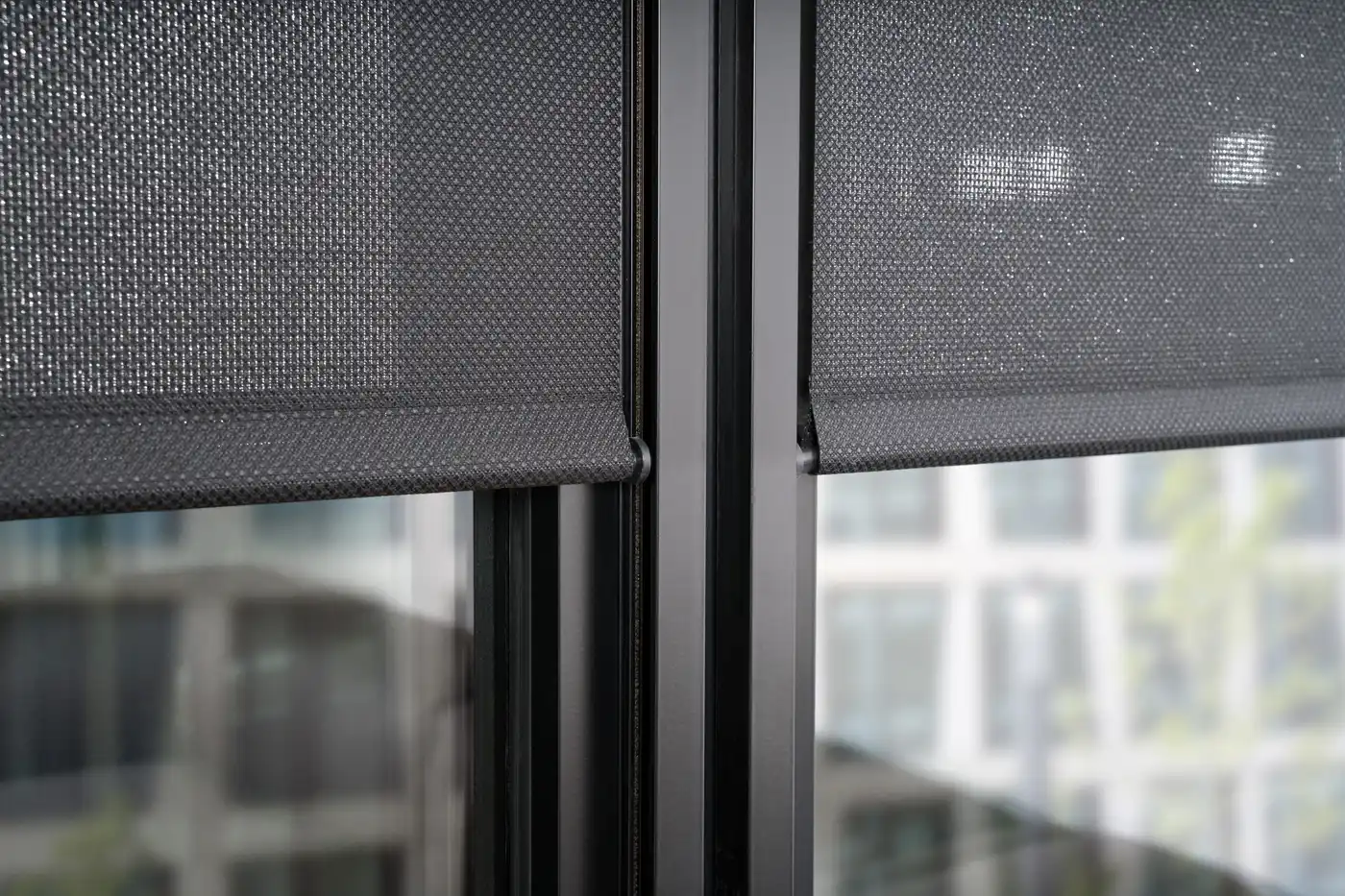Nahaufnahme zweier dunkler Screen-Rollos mit gesäumter Unterkante an einem schwarzen Fensterpfosten.