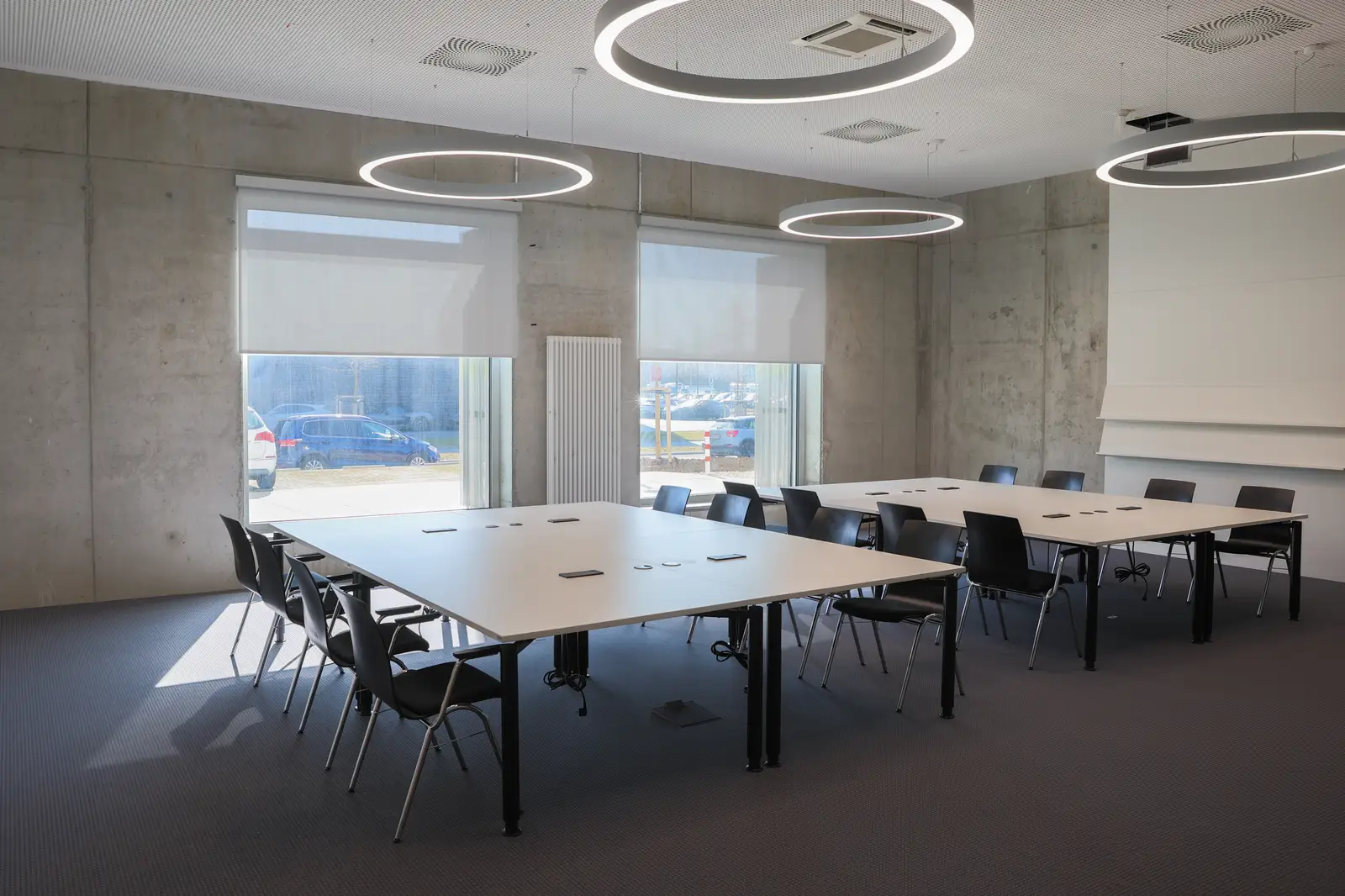 Besprechungsraum mit weißen Tischgruppen, schwarzen Stühlen und Ring-Leuchten vor Sichtbetonwand mit zwei Fenstern.