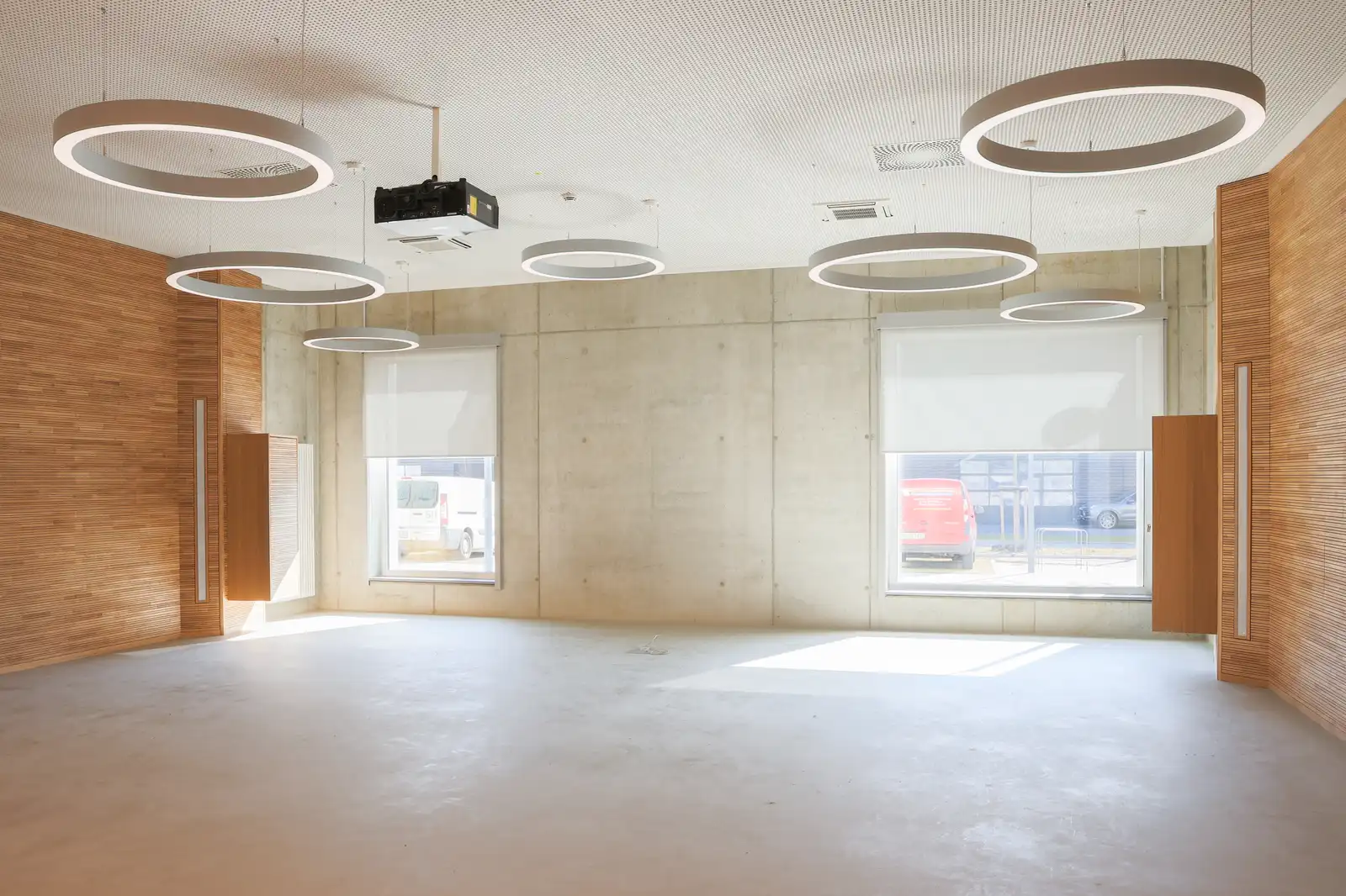 Heller, leerer Raum mit Holzverkleidung, Betonwänden, großen Fenstern und runden Deckenleuchten.
