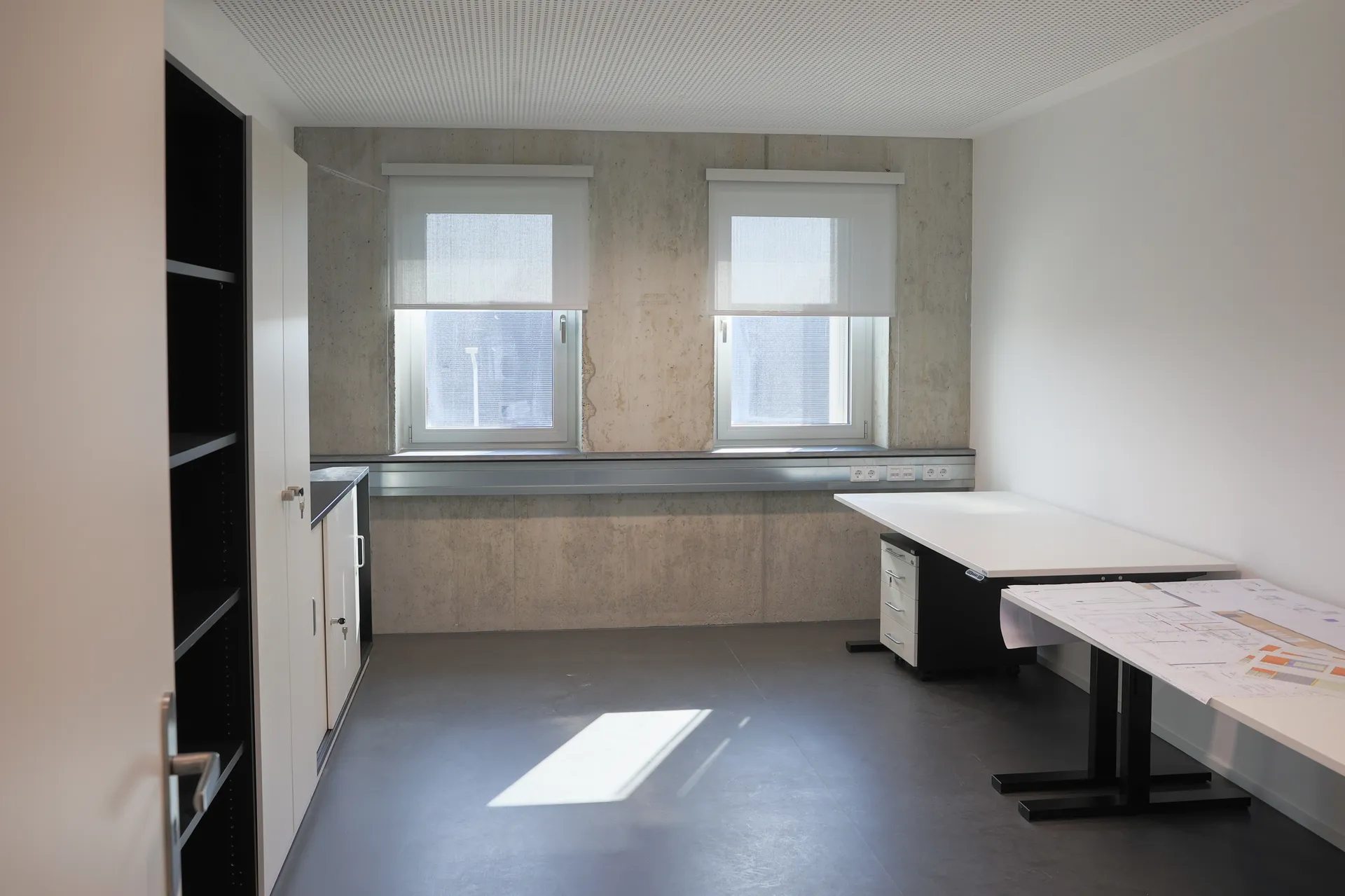Leeres Büro mit Sichtbetonwand, zwei Fenstern und gelochter Decke, rechts weiße Schreibtische mit ausgelegten Plänen.
