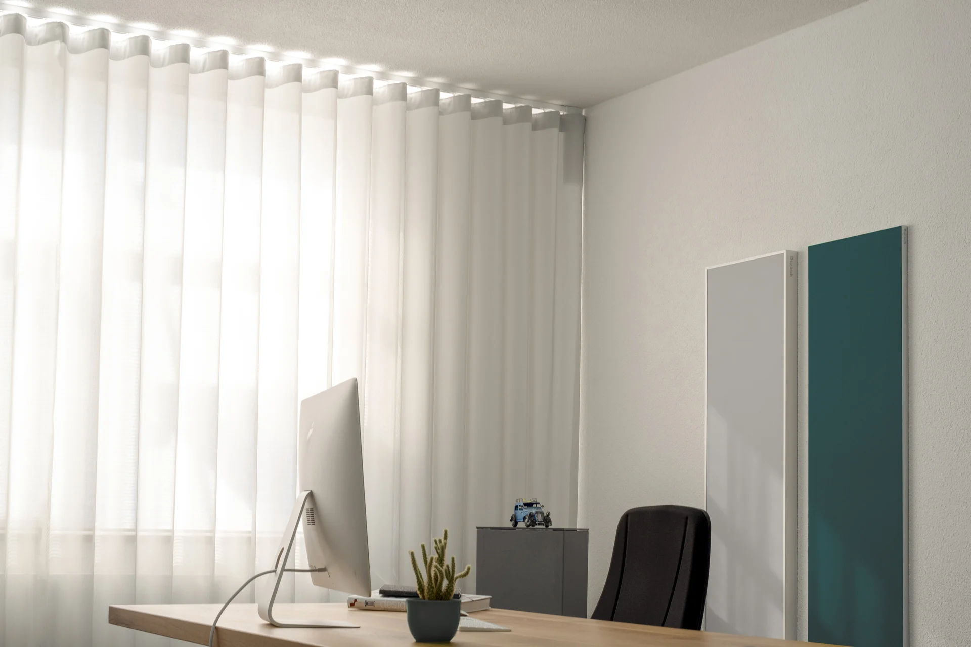 Büro mit Holzschreibtisch und raumhohem weißem Wellenvorhang, an der Wand zwei hochformatige Paneele in Weiß und Petrol.