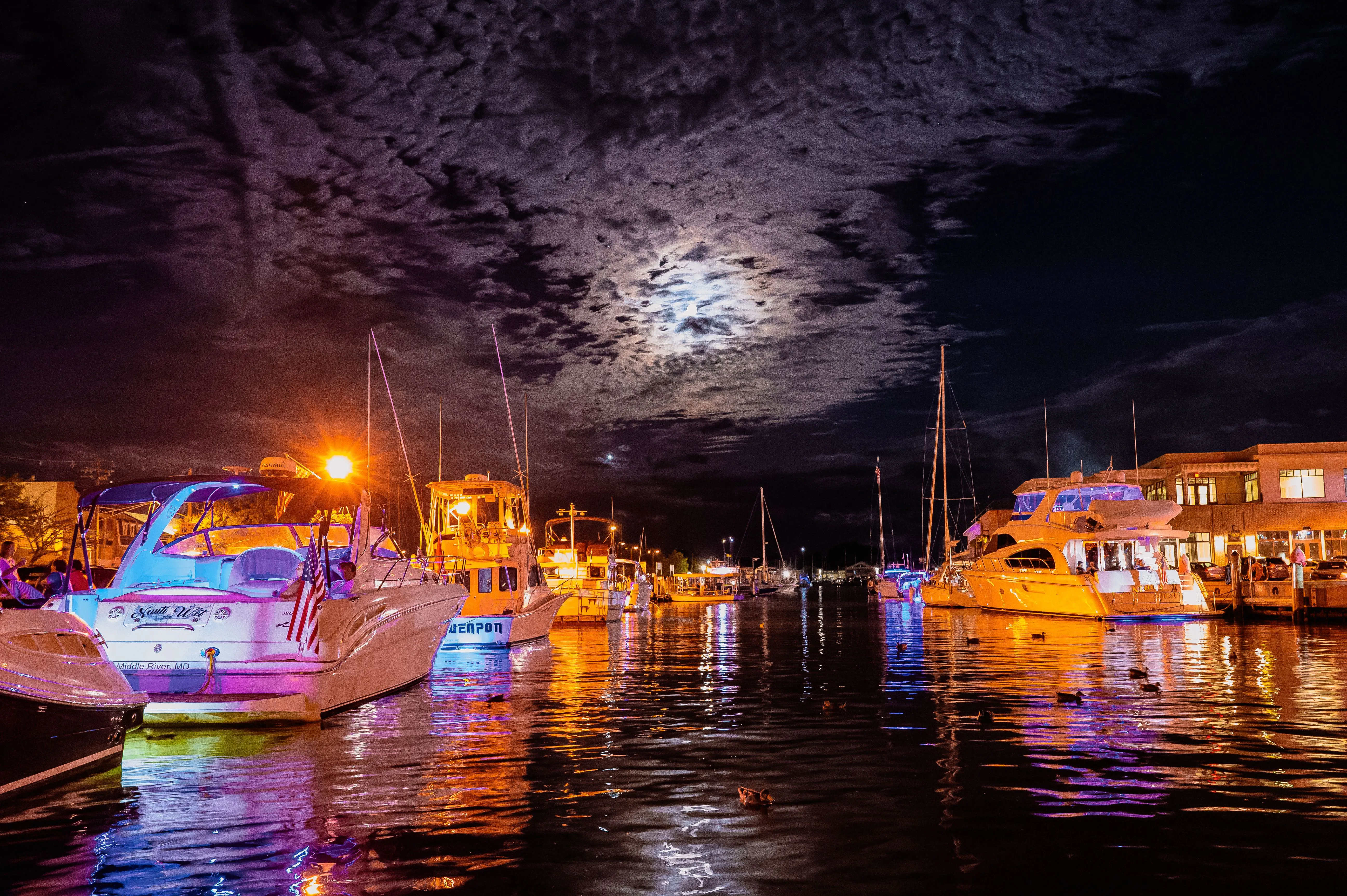 Night marina scene with docked boats, illuminated water, and a moon behind clouds.
