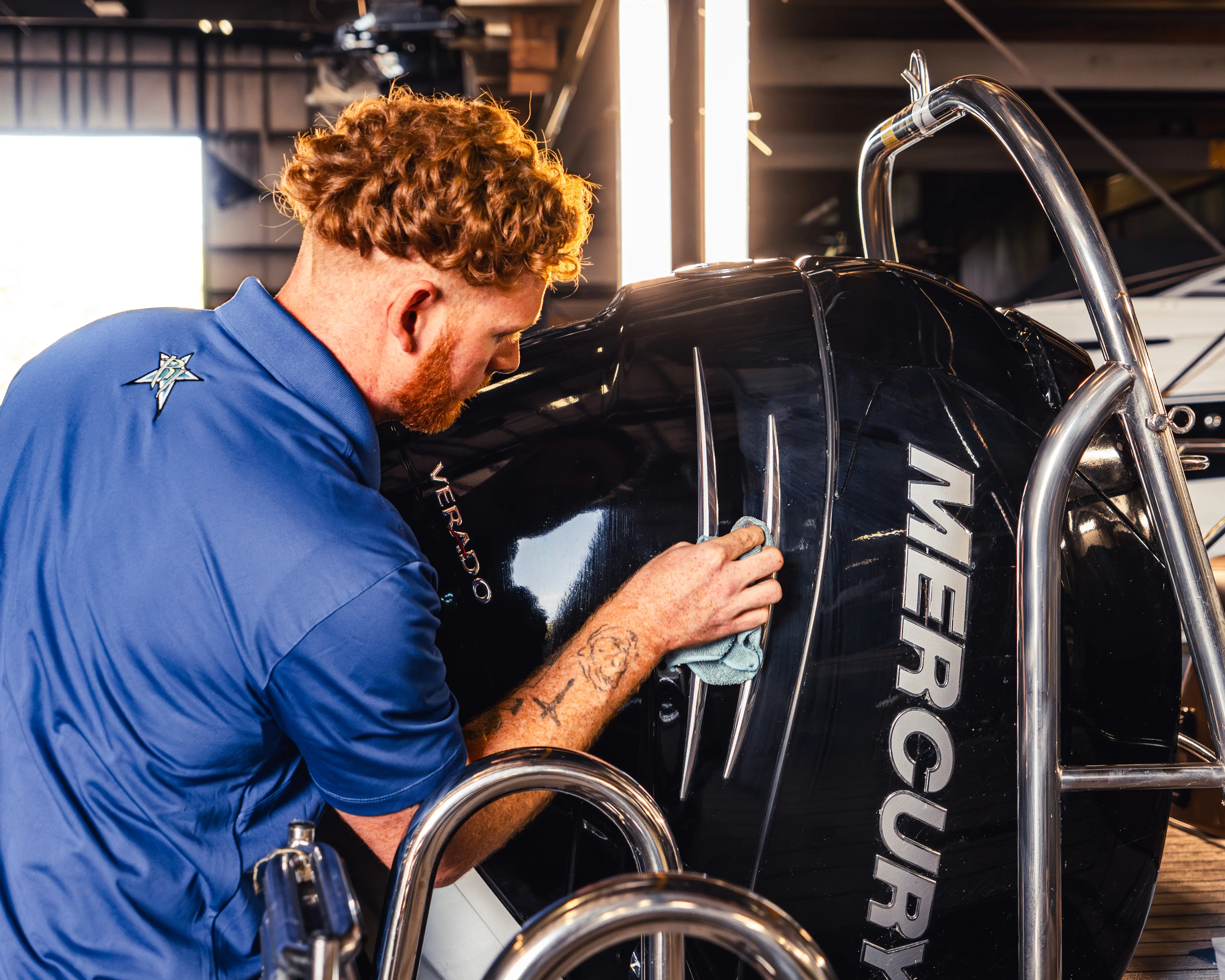 Man with red hair and tattoos polishing a black Mercury boat engine in a workshop.