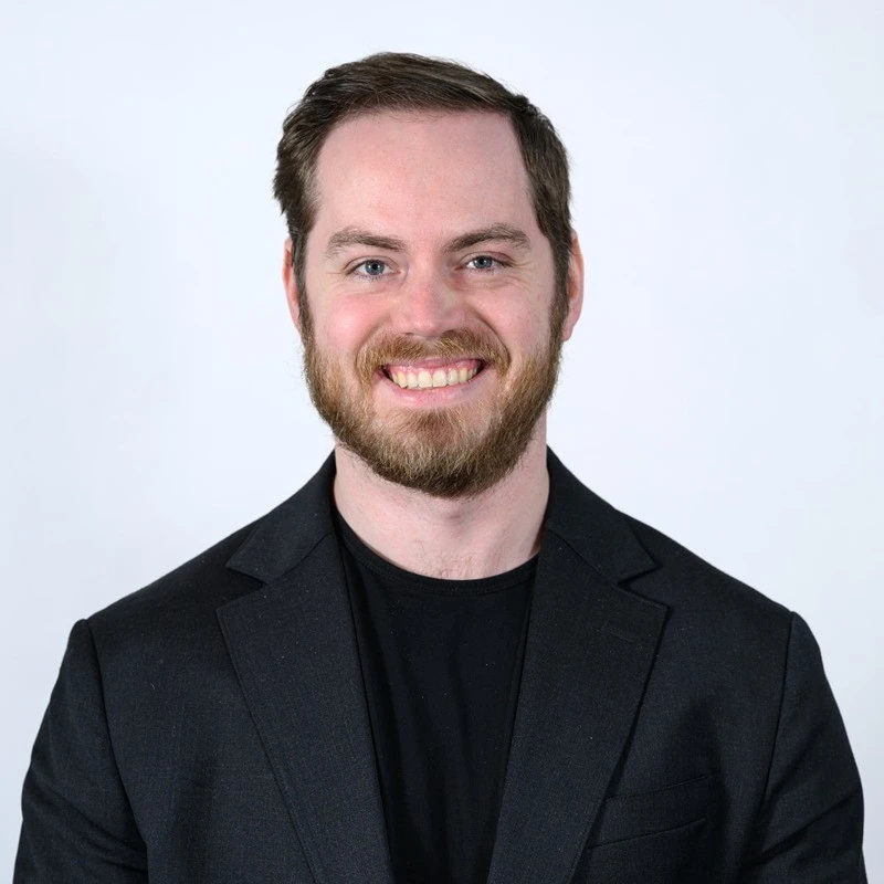 Professional headshot of a smiling man wearing a black blazer against a light background.