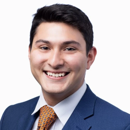 Professional headshot of a smiling man in a blue suit and orange tie against a light background.