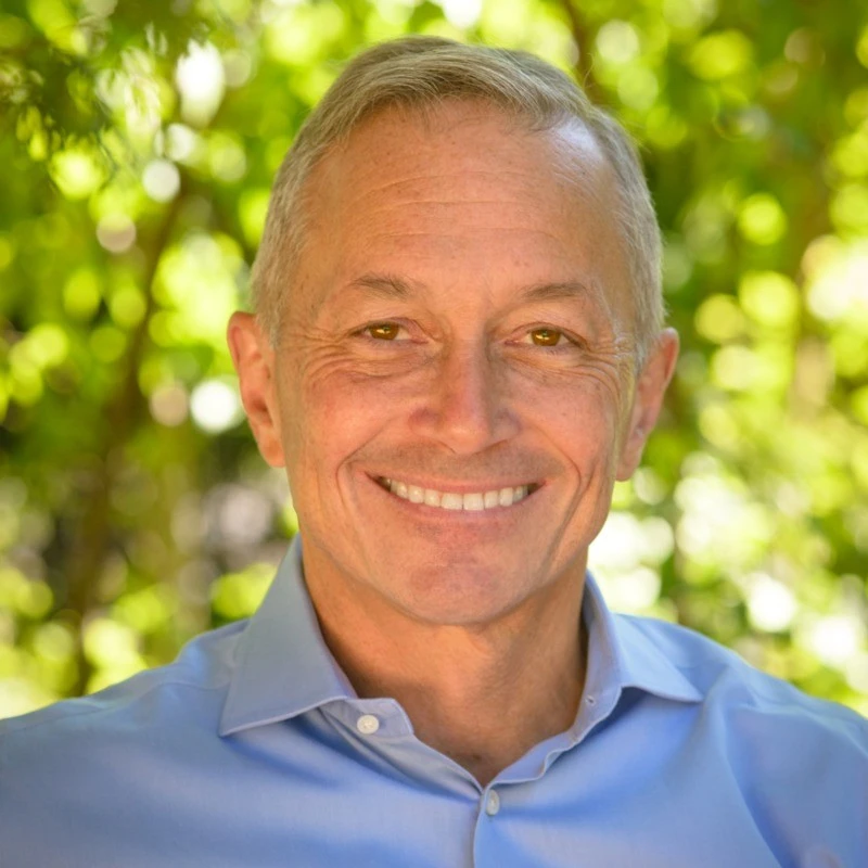 Professional headshot of a smiling man wearing a light blue shirt outdoors.