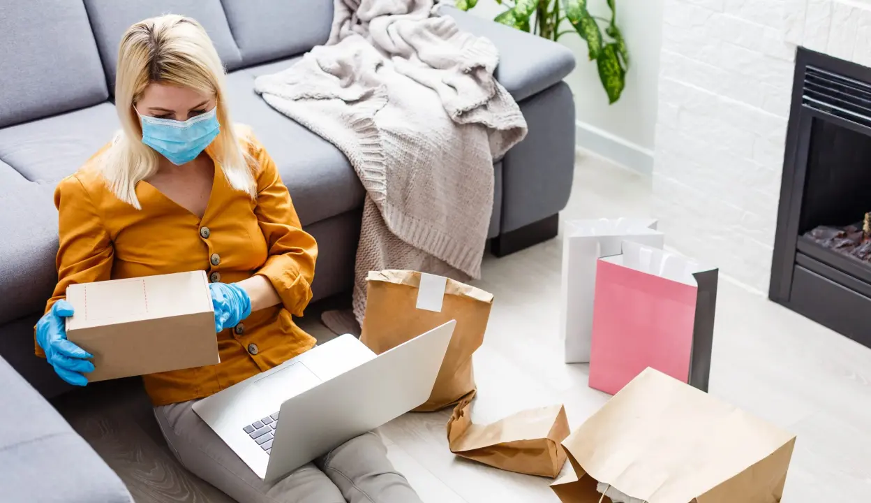 Woman wearing a face mask and gloves while reviewing a package on a laptop at home, illustrating e-commerce and online shopping behavior.