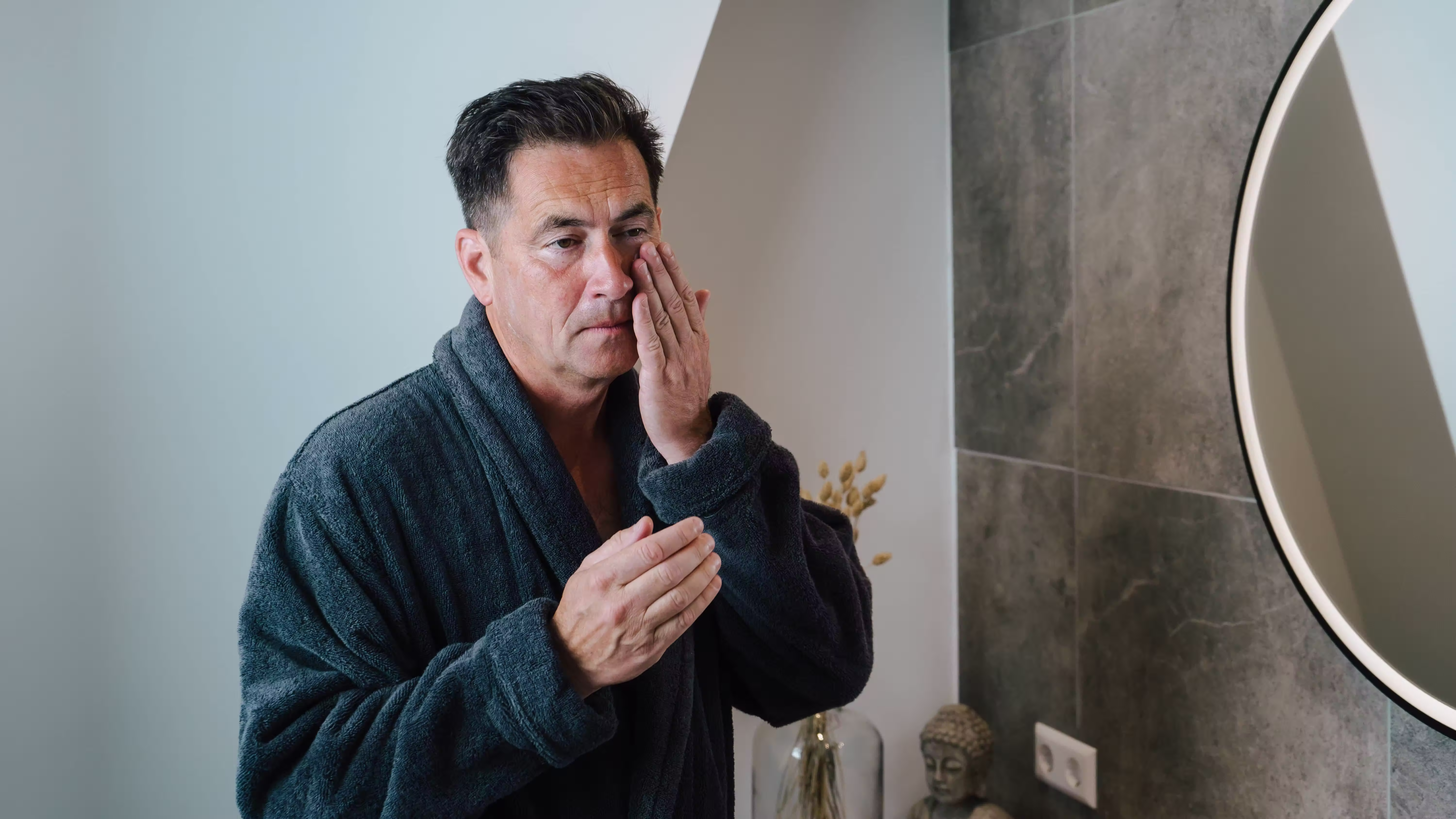 Man in a bathrobe applying skincare in front of a bathroom mirror, Olivida
