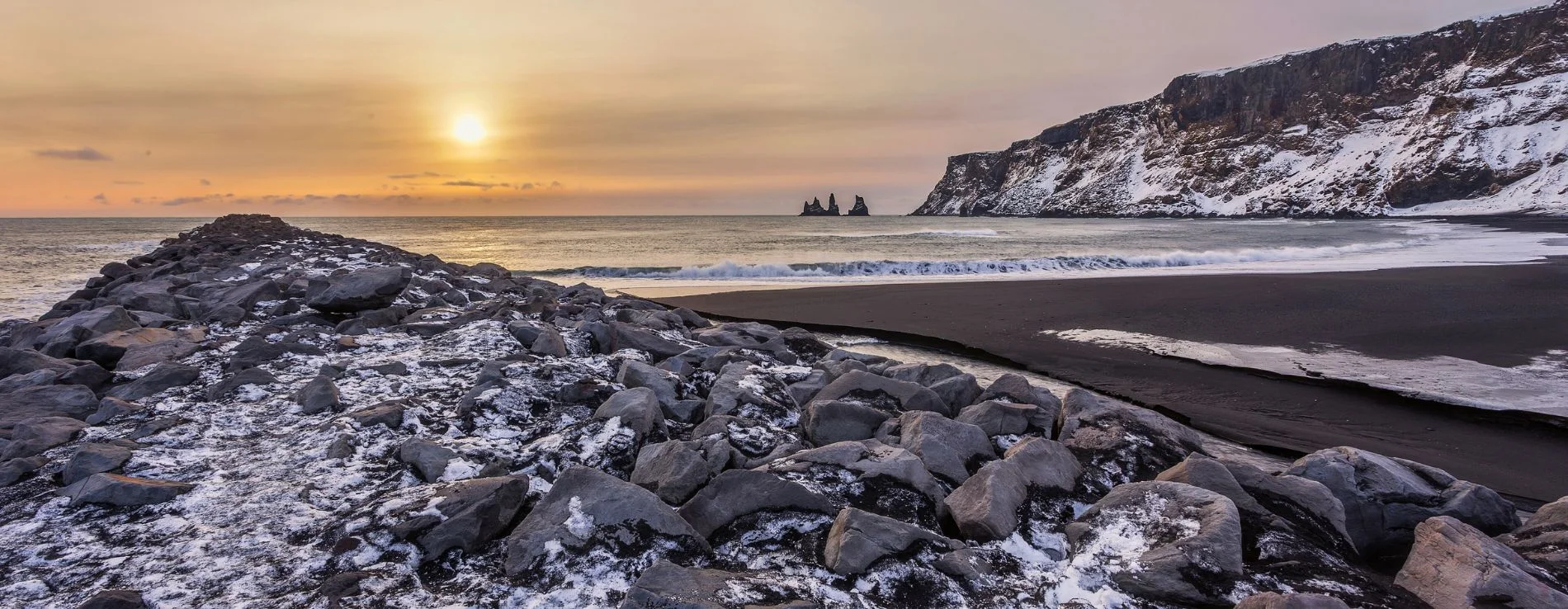Vikurfjara black sand beach in winter with light snow covering the ground and view of Reynisdrangar at sunset.