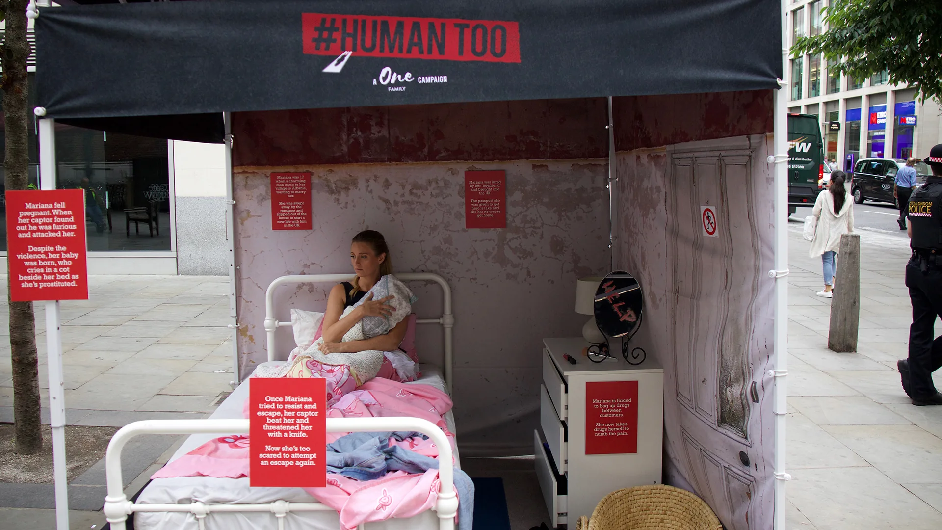 A #HUMANTOO street installation: an actor sits on a bed in a shabby bedroom set inside a gazebo, with story panels on the walls