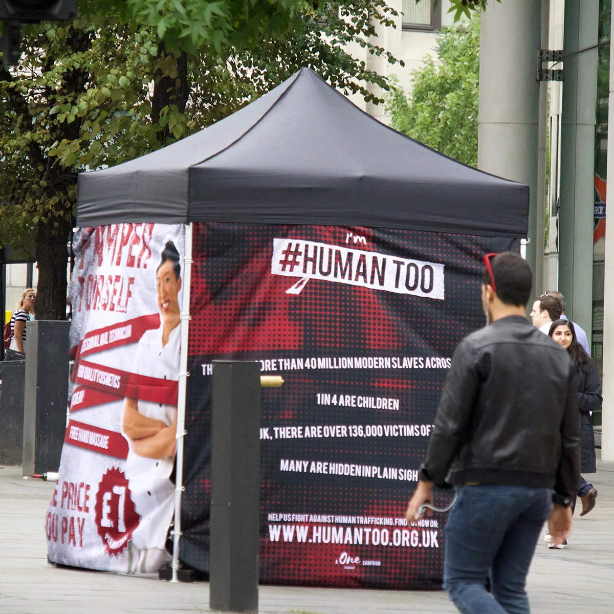 A #HUMANTOO gazebo on a city street with campaign graphics and modern slavery facts as people walk past