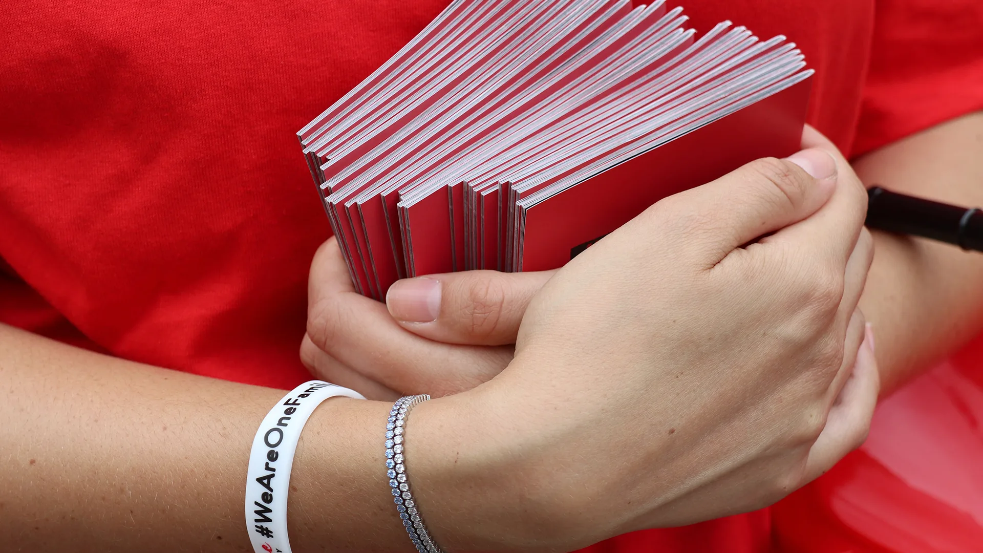 A volunteer in a red T-shirt and #WeAreOneFamily wristband holding a stack of red #HUMANTOO passports