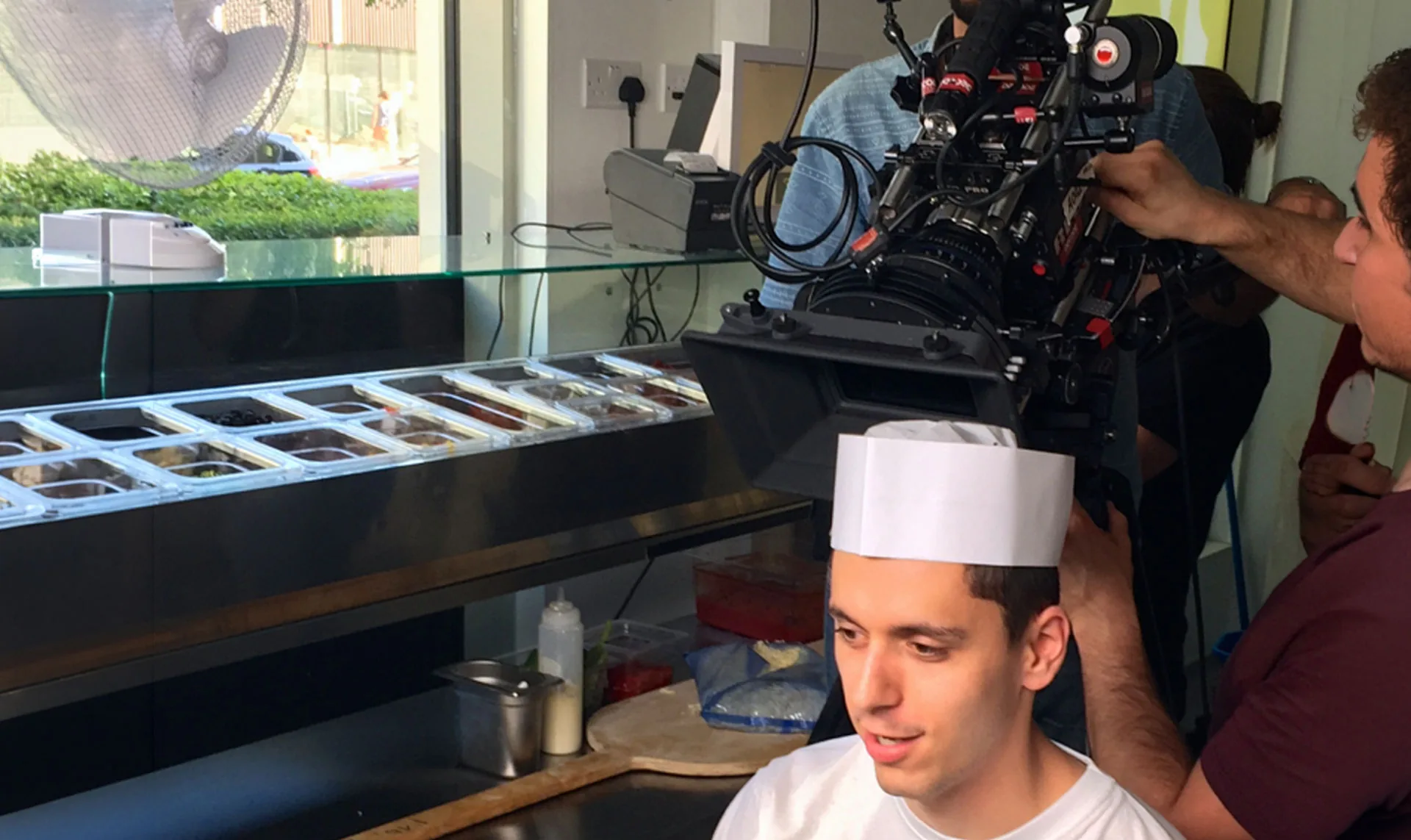 A film camera operator shooting a young chef in a white paper hat behind a Firezza counter