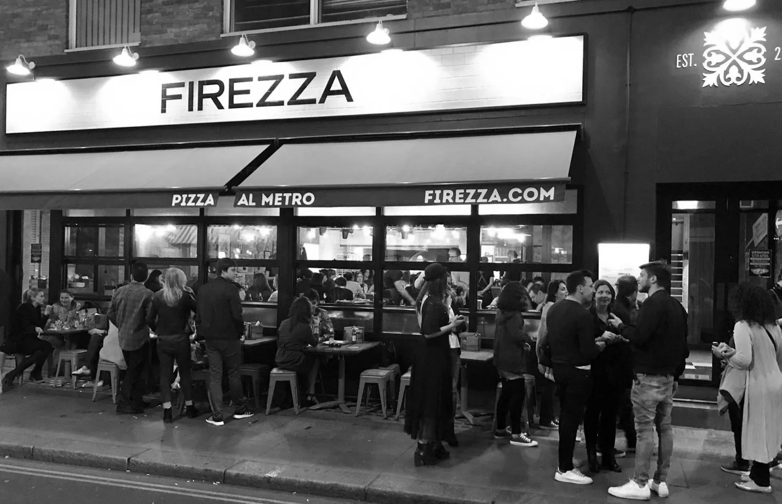 A black and white photo of the Firezza Pizza al Metro restaurant at night, with crowds outside its glass frontage