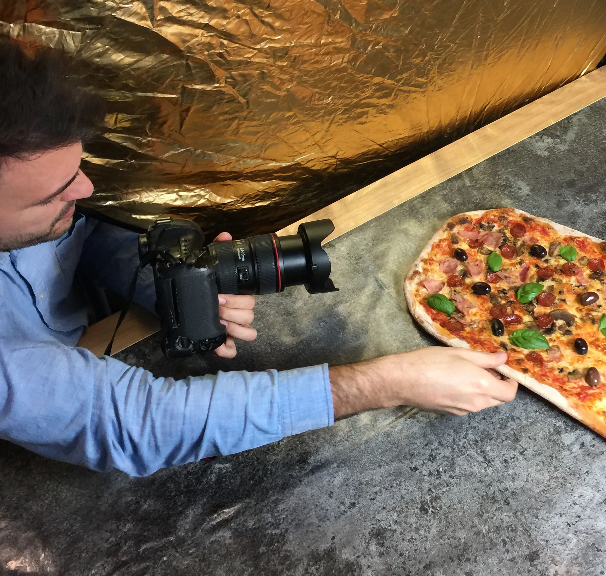 A photographer shooting a long Firezza pizza on a slate surface under gold reflector foil