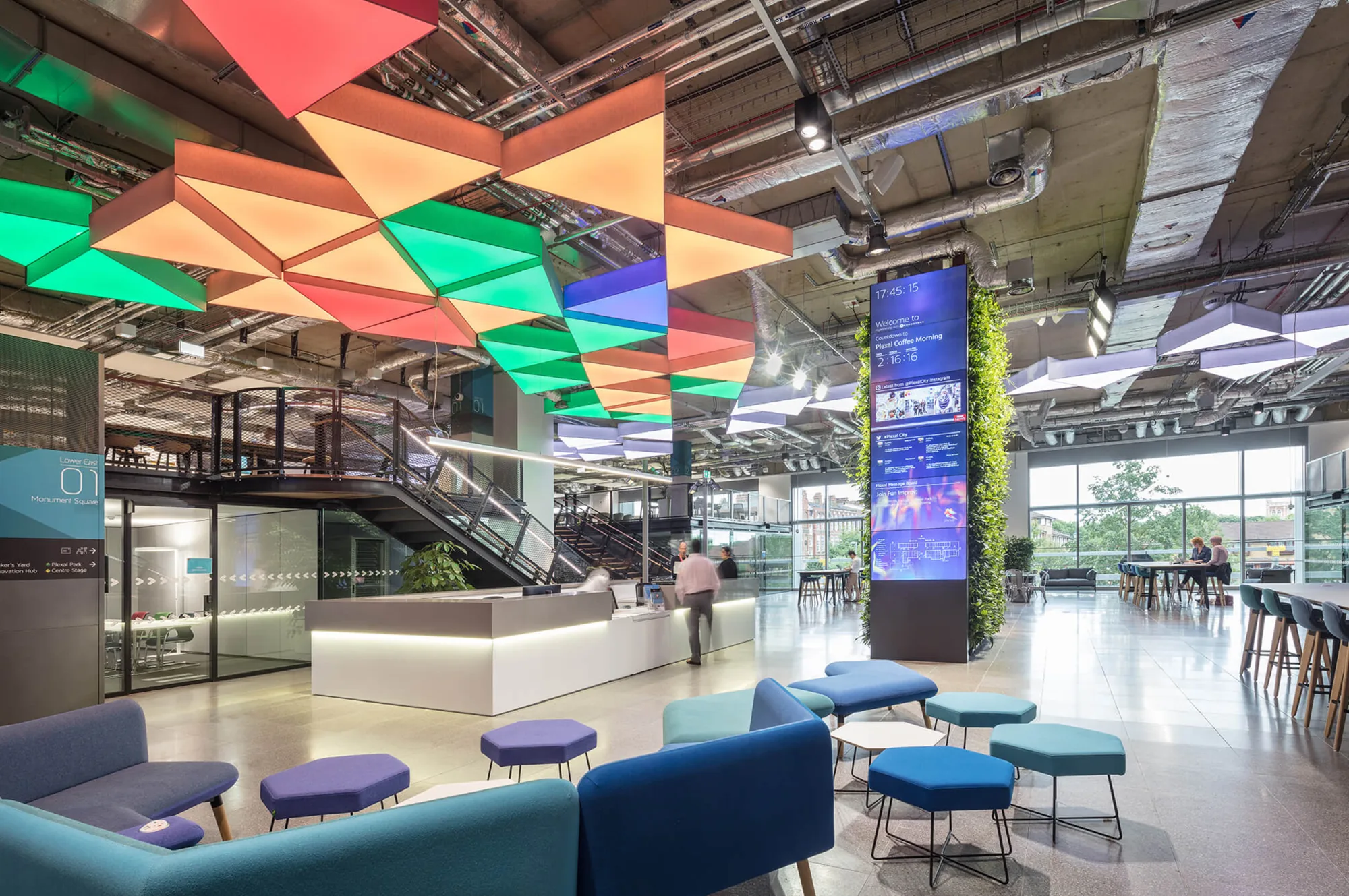 The Plexal reception and lounge, with colourful triangular ceiling panels, a digital screen tower and blue and purple seating
