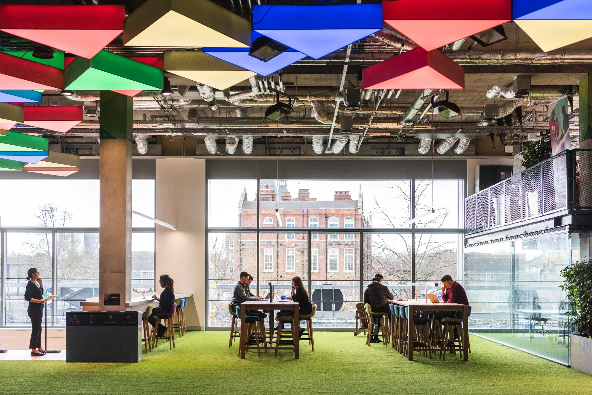 People working at tables on green carpet beneath colourful triangular ceiling lights, with windows onto the Olympic Park