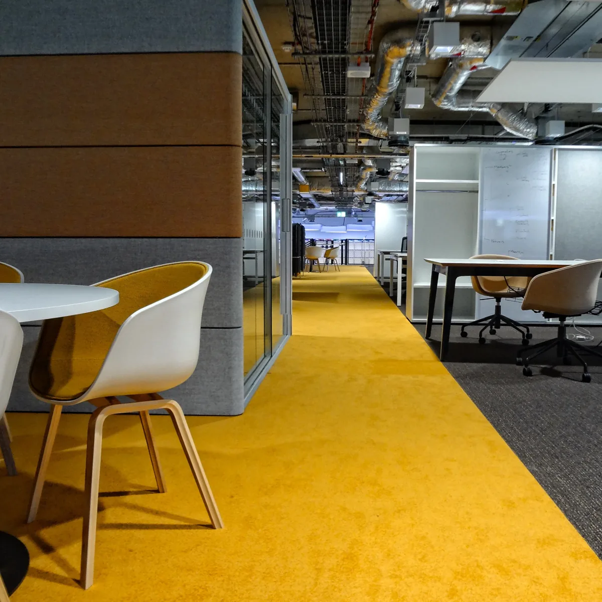 Breakout seating with white tables and mustard chairs on a bright yellow carpet path at Plexal
