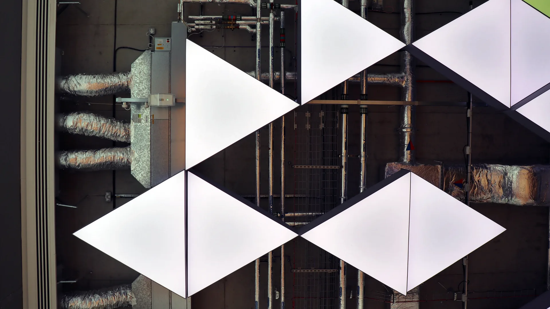 Glowing white triangular light panels against exposed ceiling pipes at Plexal