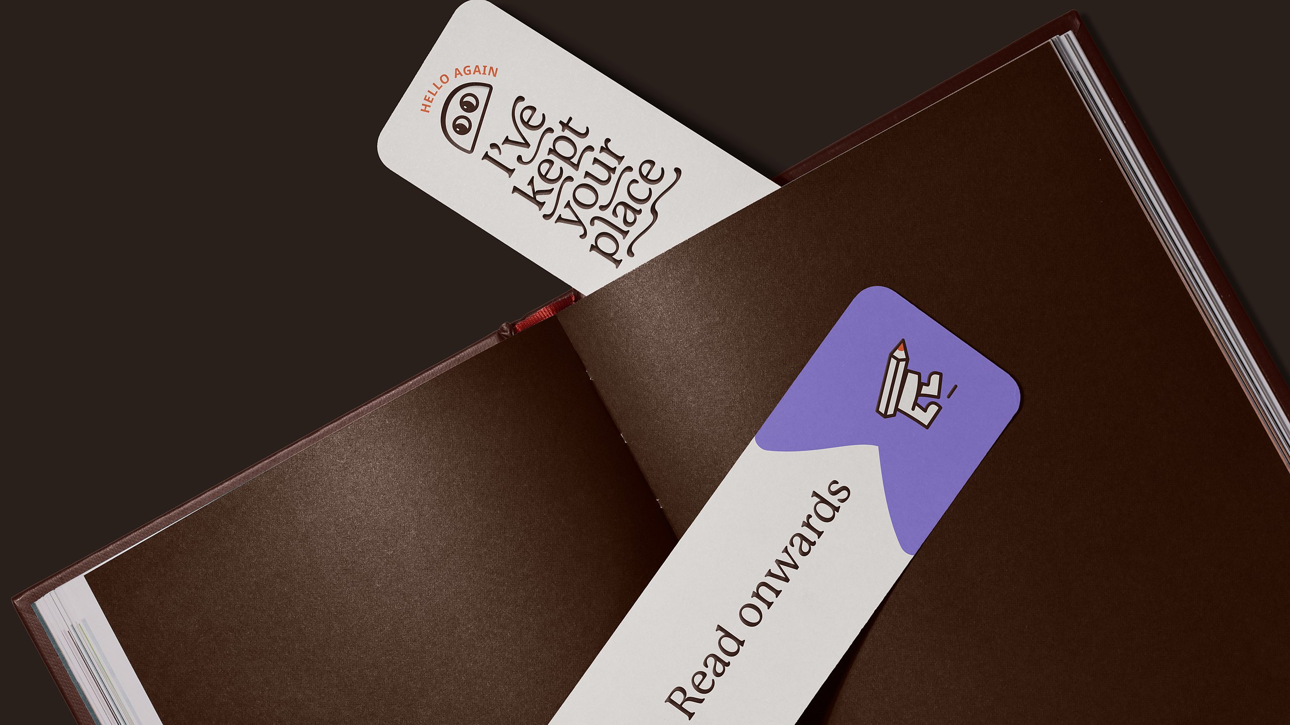 Two Libraro bookmarks in an open brown book: a cream one with Libro’s eyes and the words Hello again, I’ve kept your place, and a purple and cream one reading Read onwards with a small Libro icon