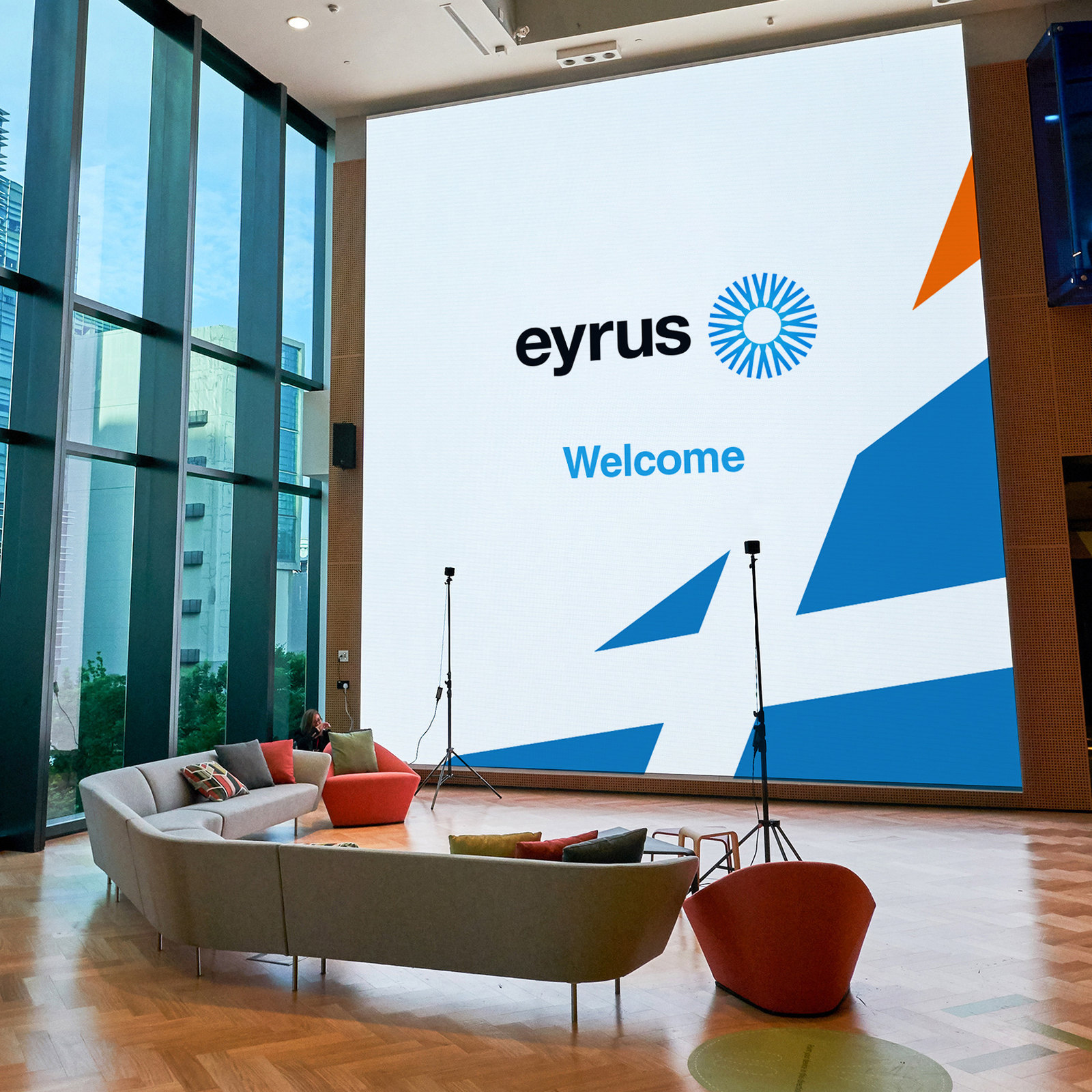 A large screen in a glass-walled lobby showing the Eyrus logo and the word Welcome over blue and orange shards from the brand’s X shape