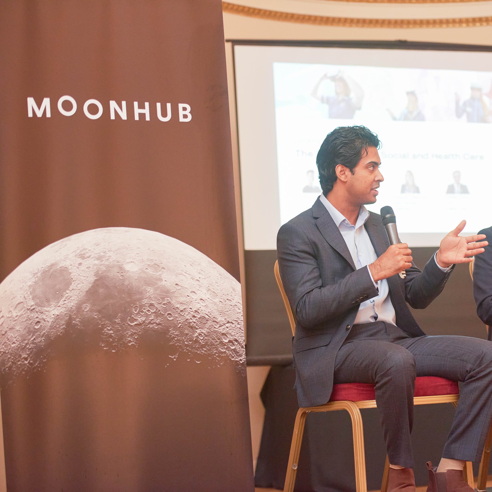 A speaker with a microphone on the panel at Moonhub’s dementia care event, beside a MOONHUB banner showing the moon
