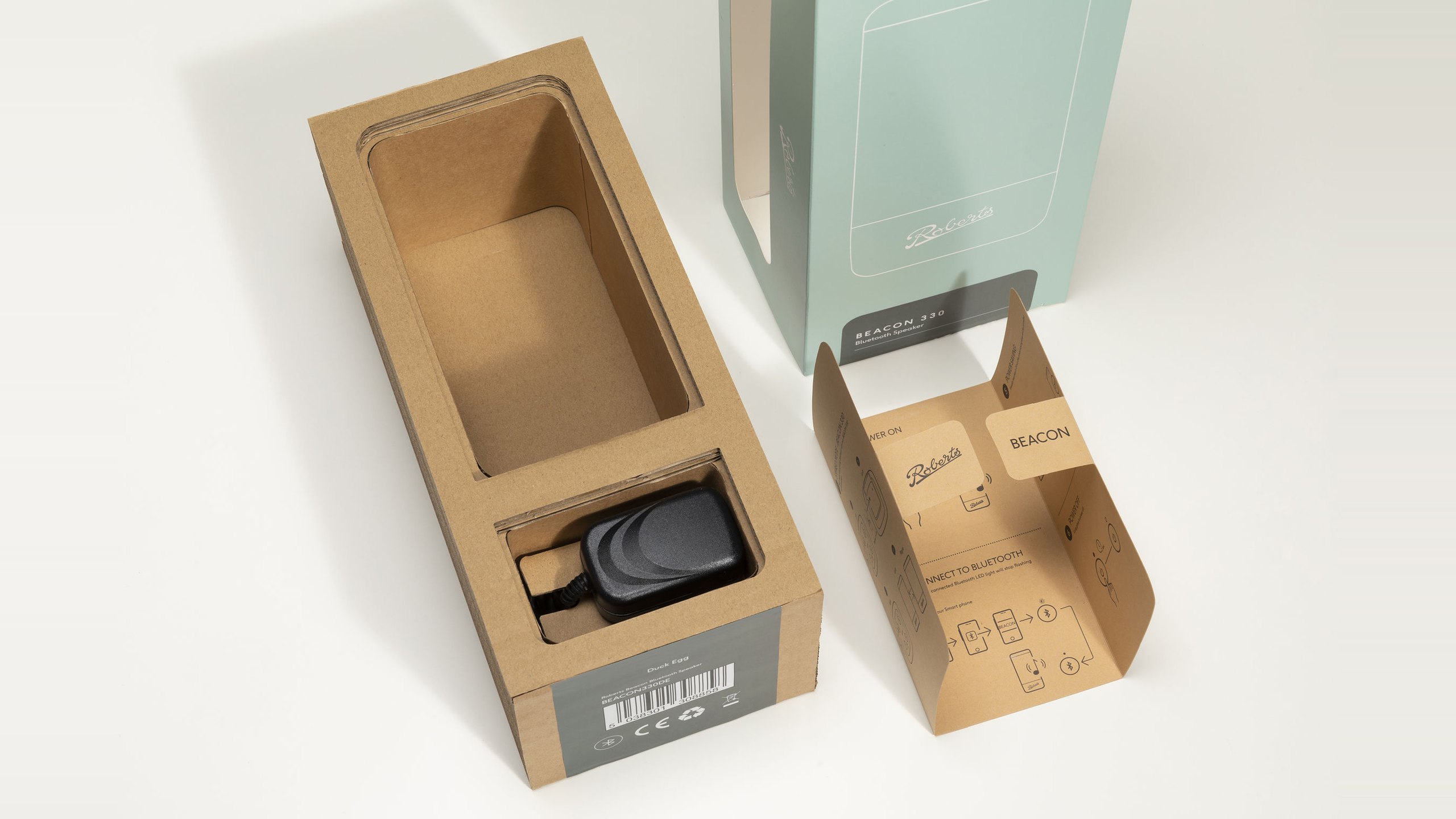 The finished Roberts Beacon 330 pack in Duck Egg, unboxed: a layered corrugated card tray with the speaker recess empty and the charger in its own compartment, a Duck Egg label on the end, the pale green sleeve with its side window and a kraft quick start insert printed with Roberts and BEACON