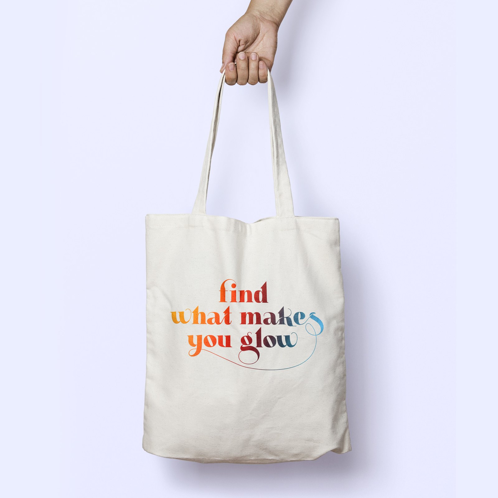 A hand holding a canvas tote bag printed with find what makes you glow in orange and blue lettering