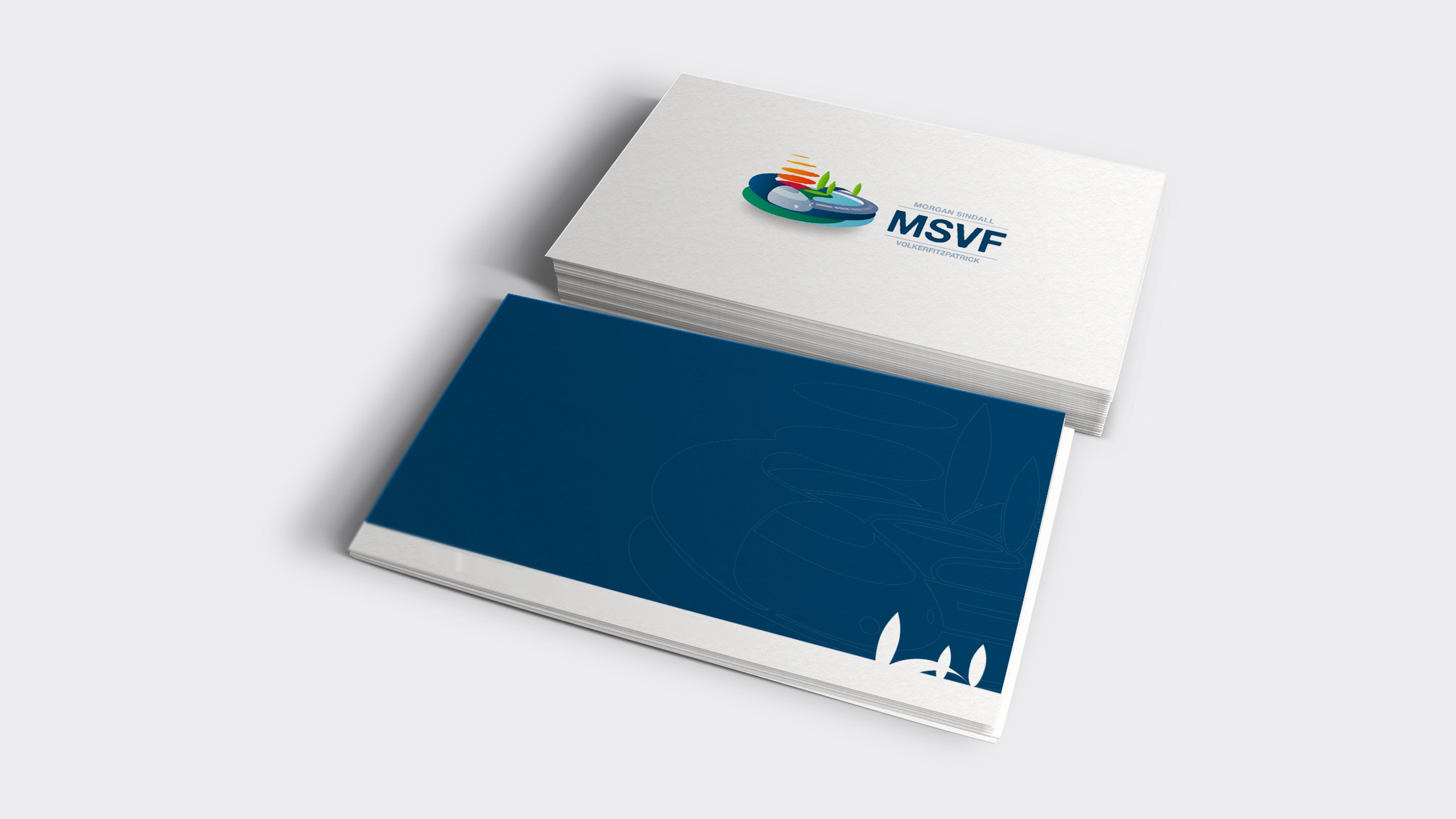 A stack of MSVF business cards on a pale grey surface: the white front carries the full-colour MSVF logo, and the navy back shows a faint outline of the mark with white tree silhouettes in the corner