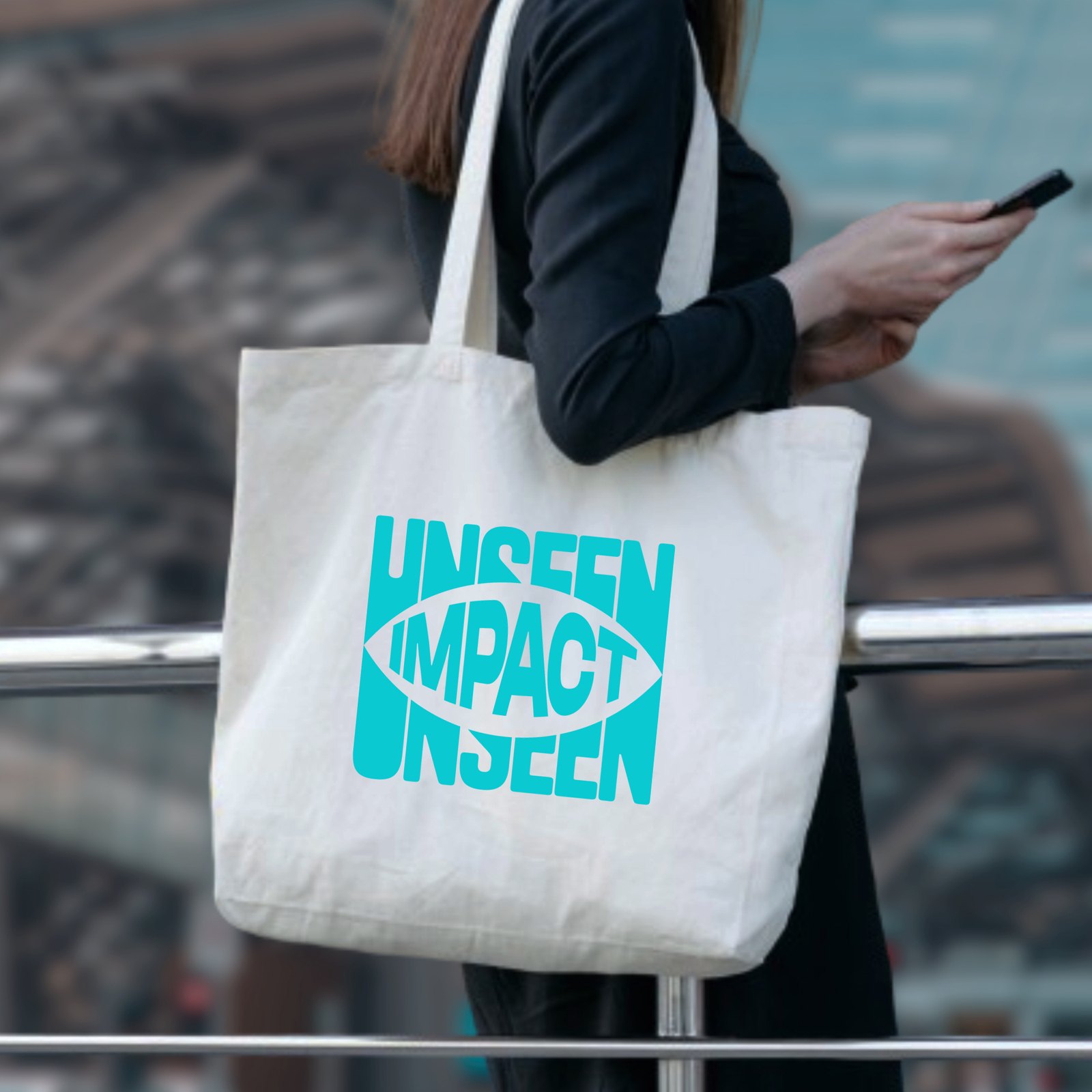 A canvas tote bag carried over the shoulder, printed with the UNSEEN IMPACT eye logo in cyan