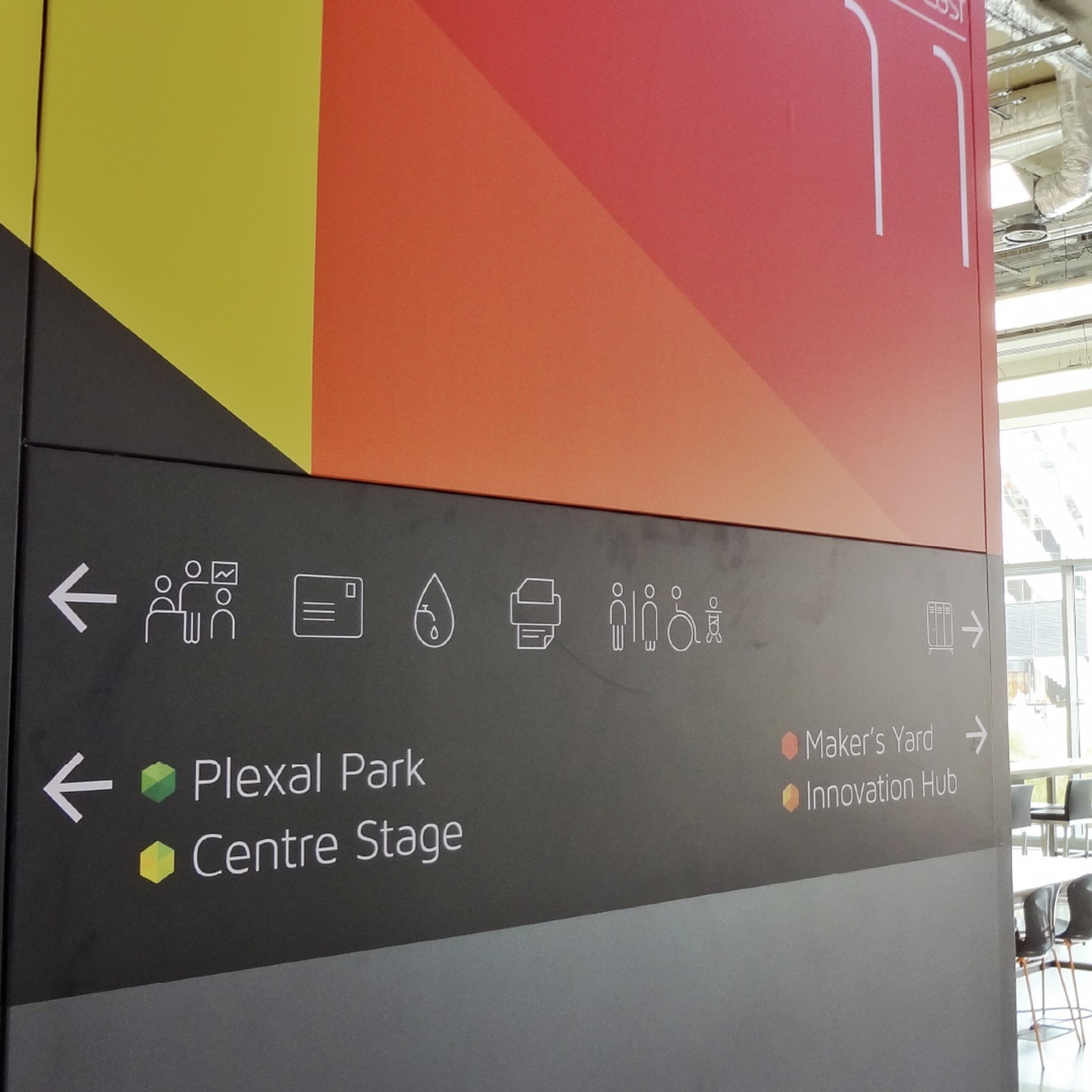 A Plexal wayfinding wall with bold red, orange and yellow colour blocks above icons and directions to Plexal Park and Centre Stage
