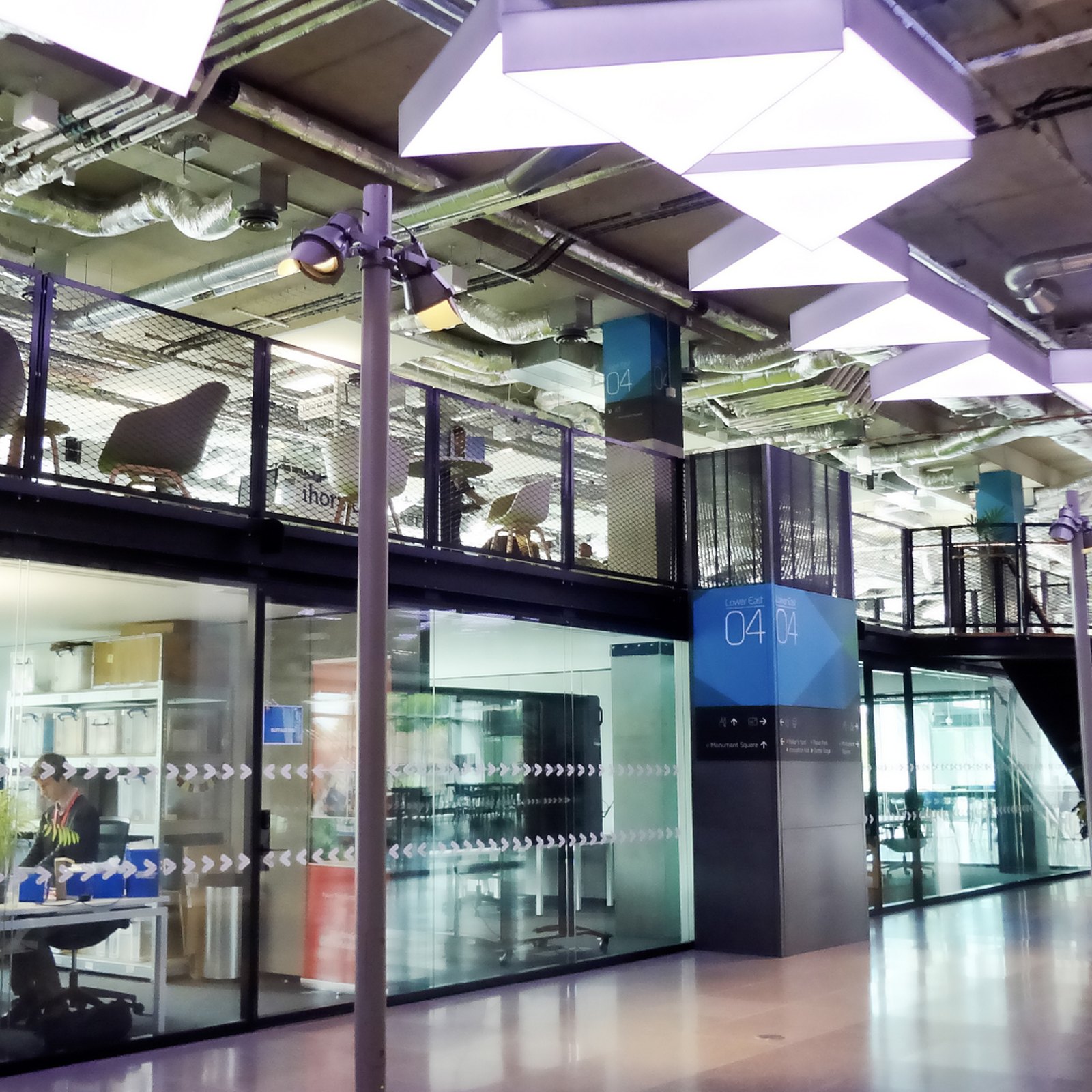 Glass-fronted studios and a mezzanine at Plexal beneath white triangular ceiling lights, with a blue wayfinding column