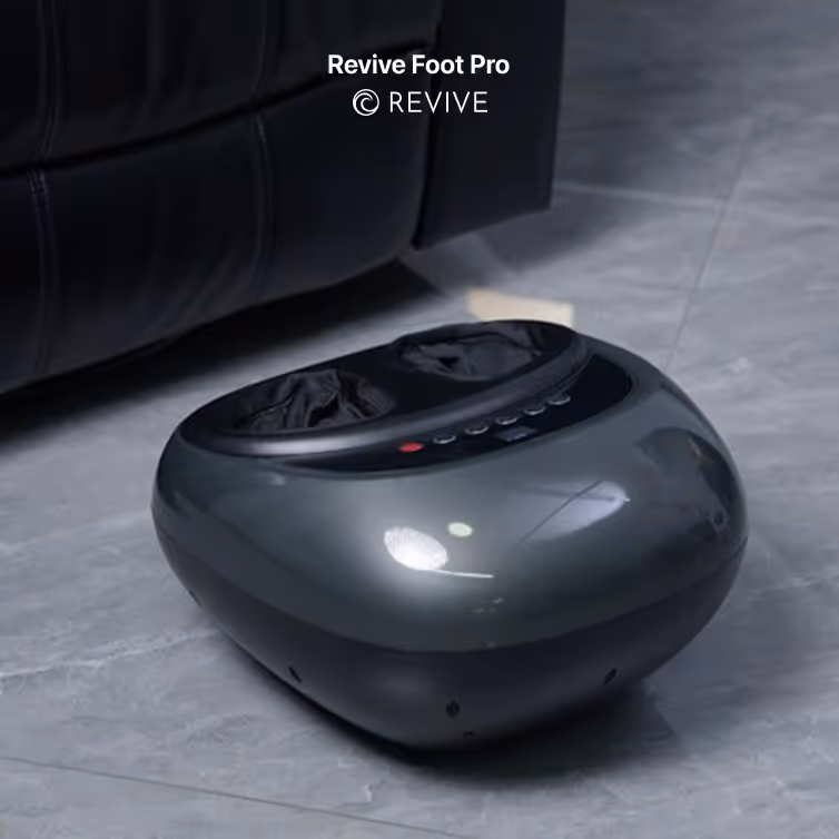 Revive Foot Pro black foot massager placed on a gray tiled floor near a black leather couch.