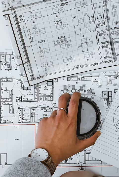 Mano con reloj sostiene un vaso sobre planos arquitectónicos junto a una libreta con notas.