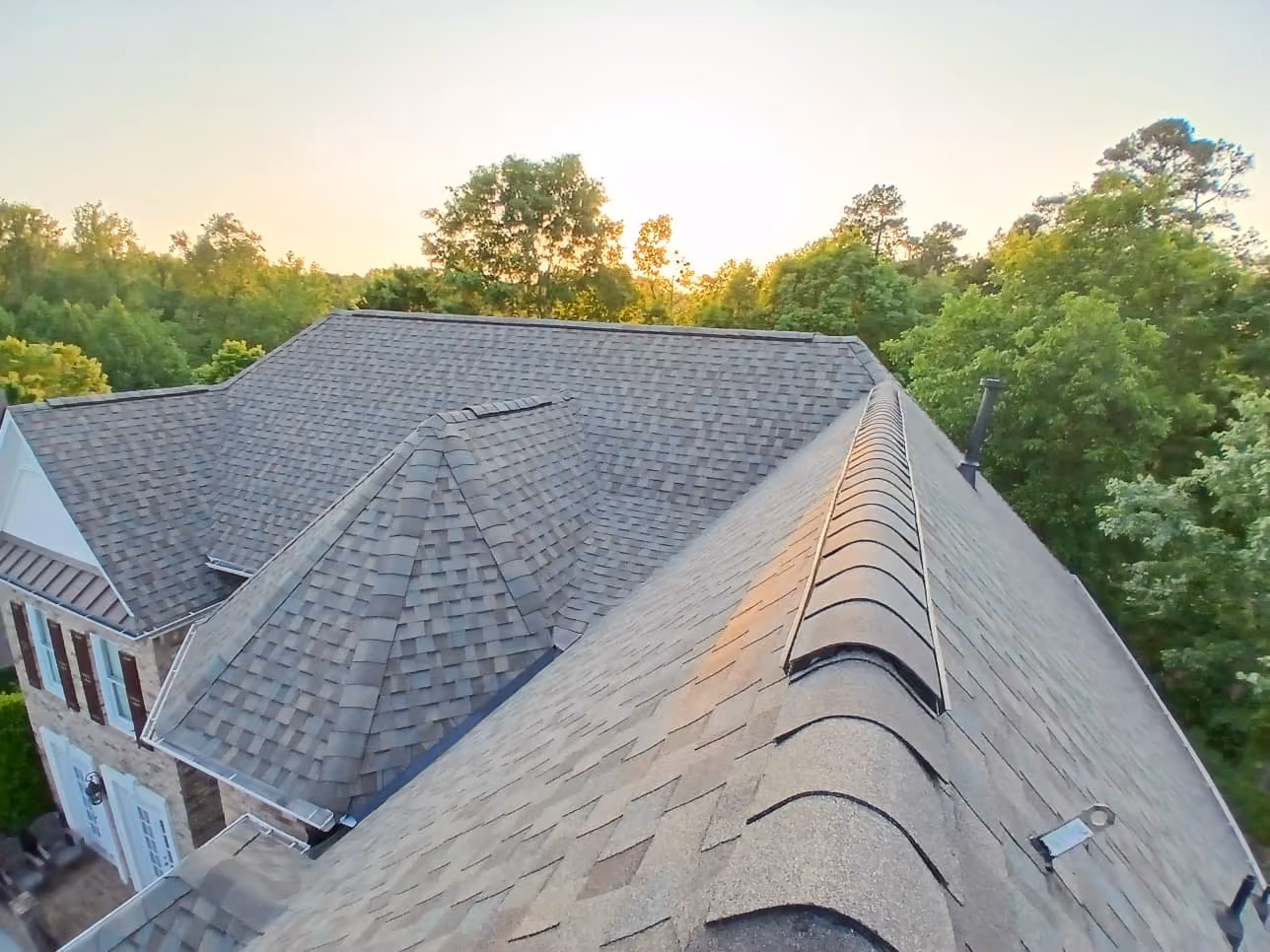 Top view of a house roof with gray shingles surrounded by green trees at sunset.